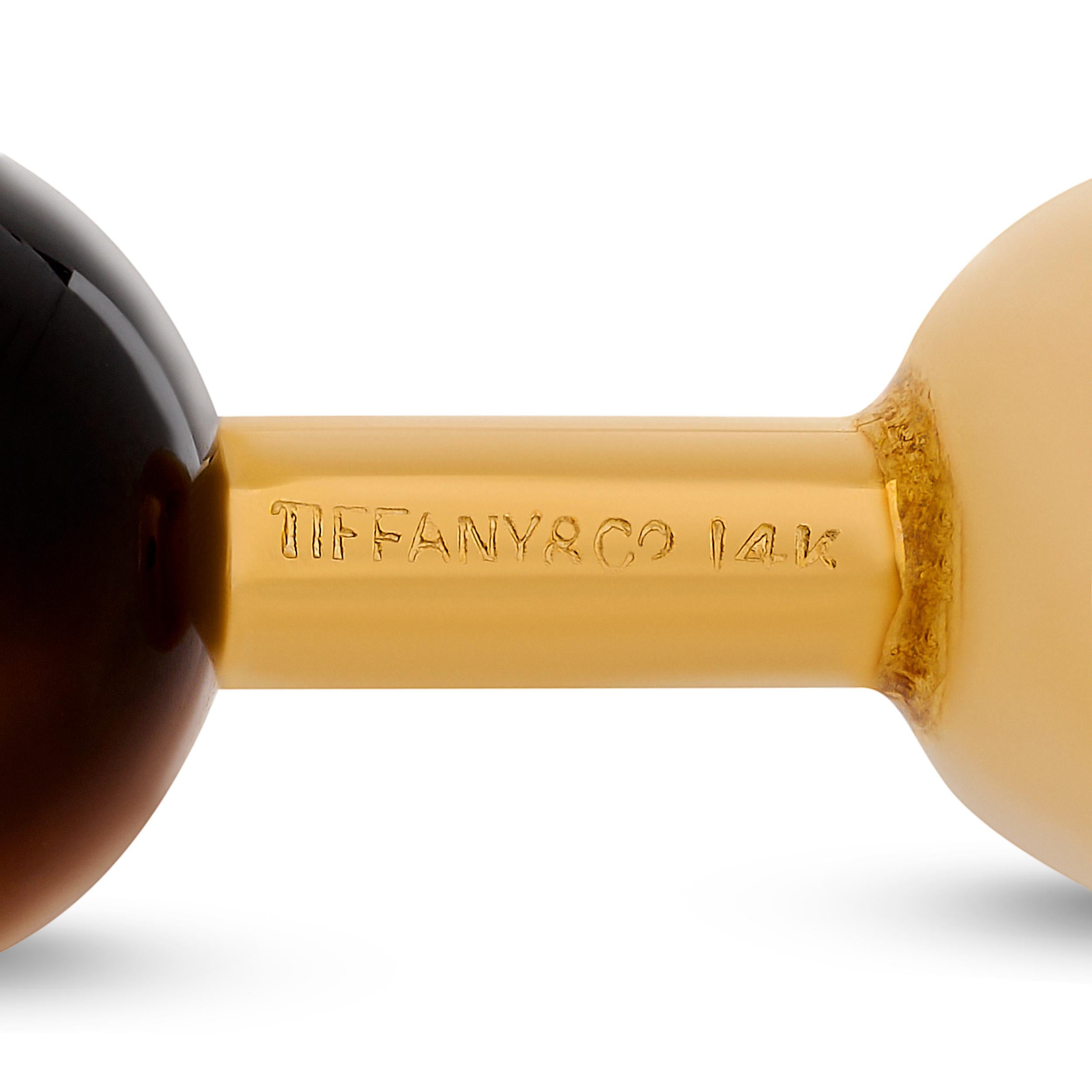 Fabriquée par Tiffany & Co, cette paire de boutons de manchette distinguée présente d'étonnantes pierres sphériques Œil de tigre élégamment serties dans des montures en or jaune 14 carats poli, dégageant un charme raffiné et sophistiqué. 


