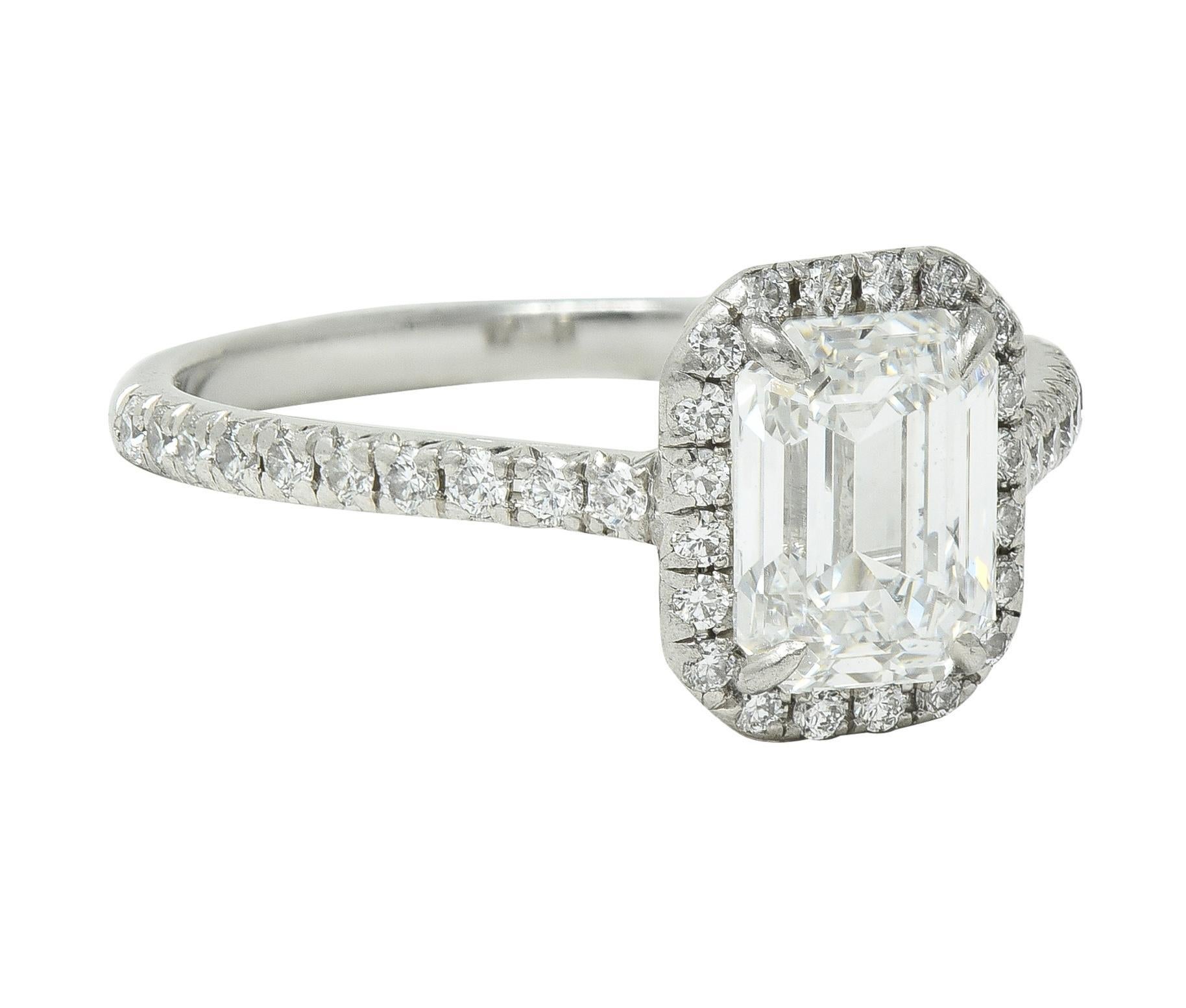 Centering an emerald cut diamond weighing 1.32 carats - F color with VS2 clarity
Prong set with a halo surround of prong set round brilliant cut diamonds
With additional diamonds prong set down the shoulders
Weighing approximately 0.27 carat total -