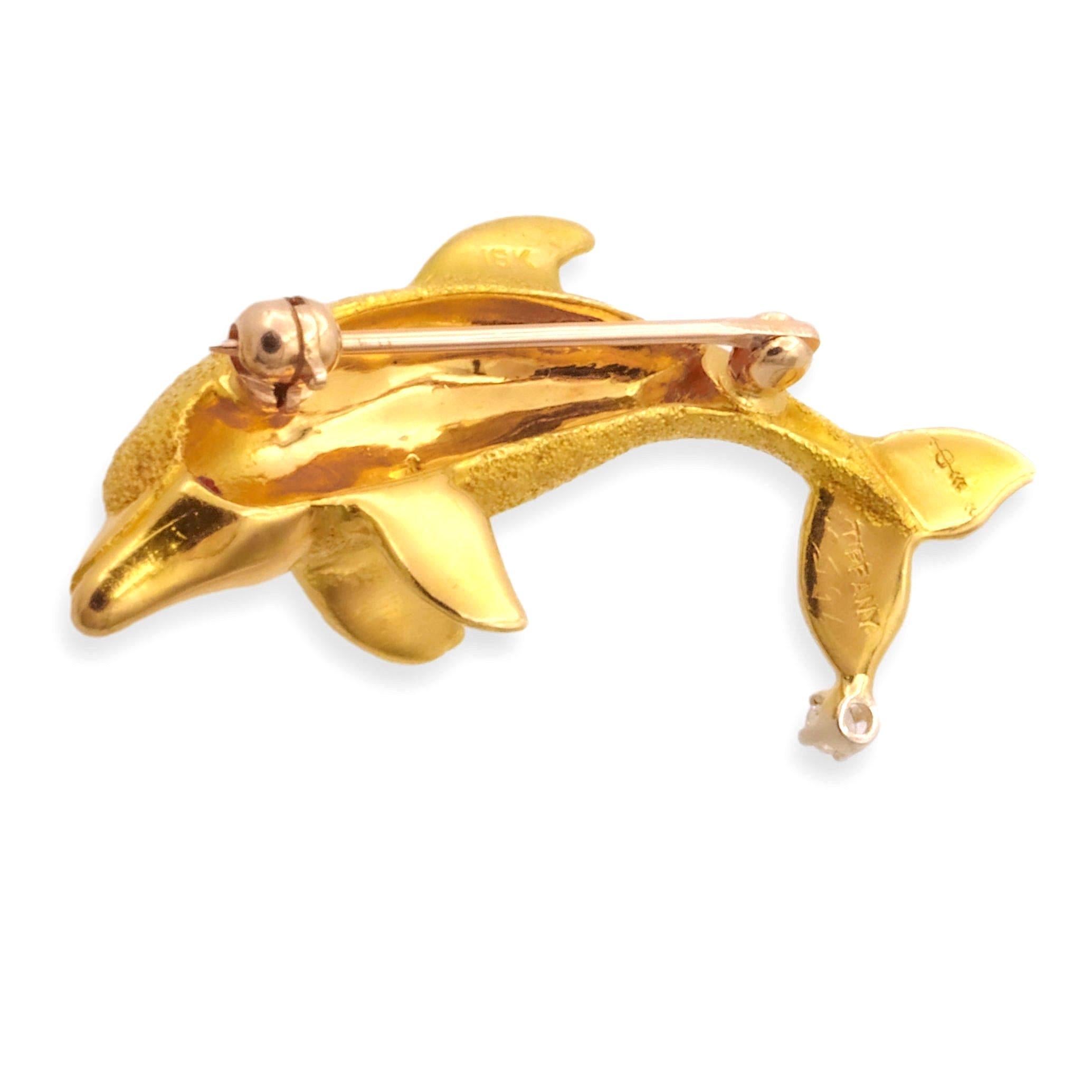 Vintage Tiffany & Co. dolphin brooch finely crafted in highly textured 18 Karat yellow gold with one deep red ruby eye and one diamond set in 4 prongs in the tail. This brooch has a sandblasted finish and texture. Round brilliant diamond weighs 0.05