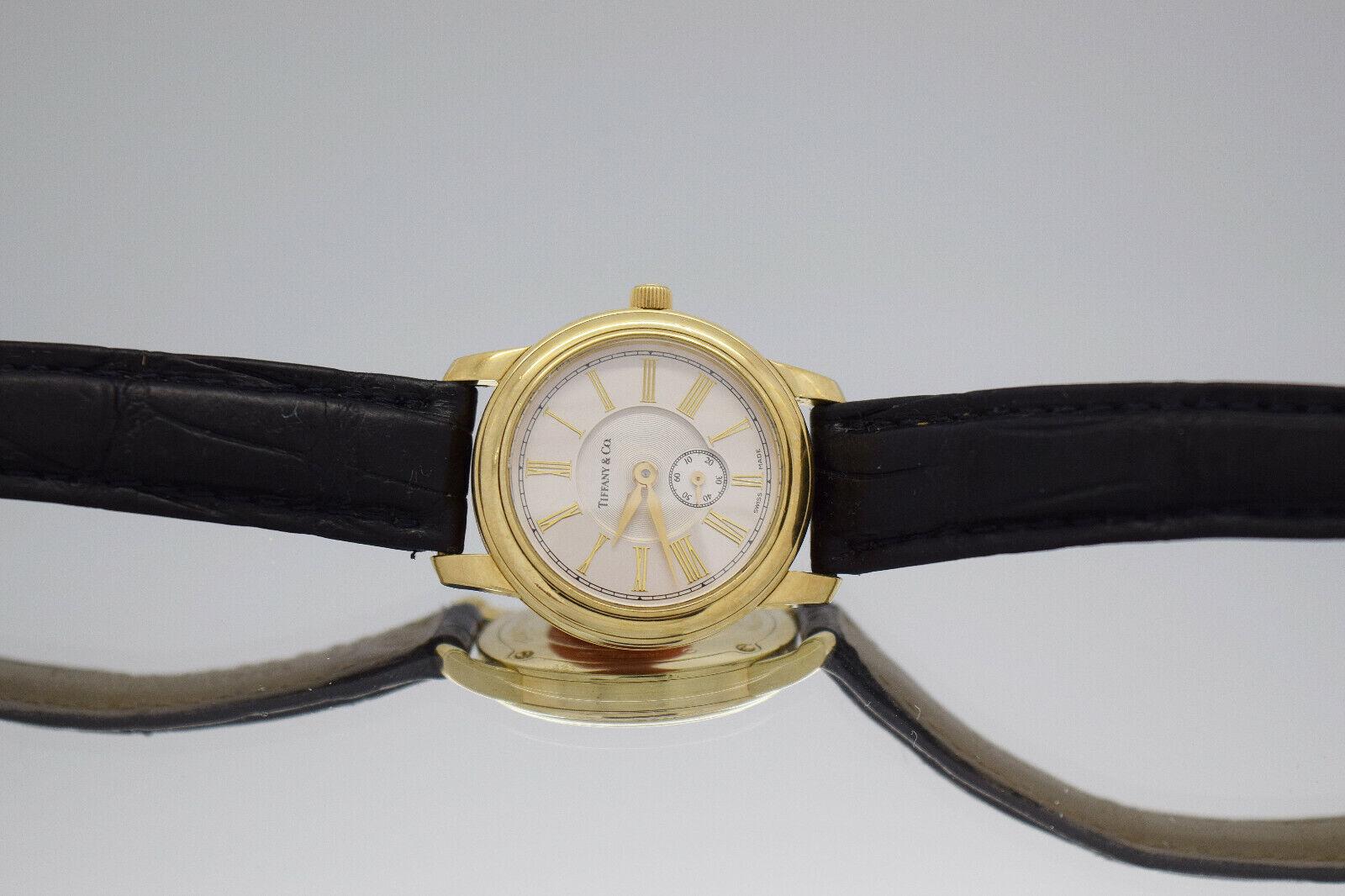 This Tiffany & Co. 18k Yellow Gold Quartz Watch is a refined timepiece featuring a 27mm round case crafted in solid yellow gold. The classic Roman numeral dial offers a timeless aesthetic, accentuated by a central guilloché-style texture and a