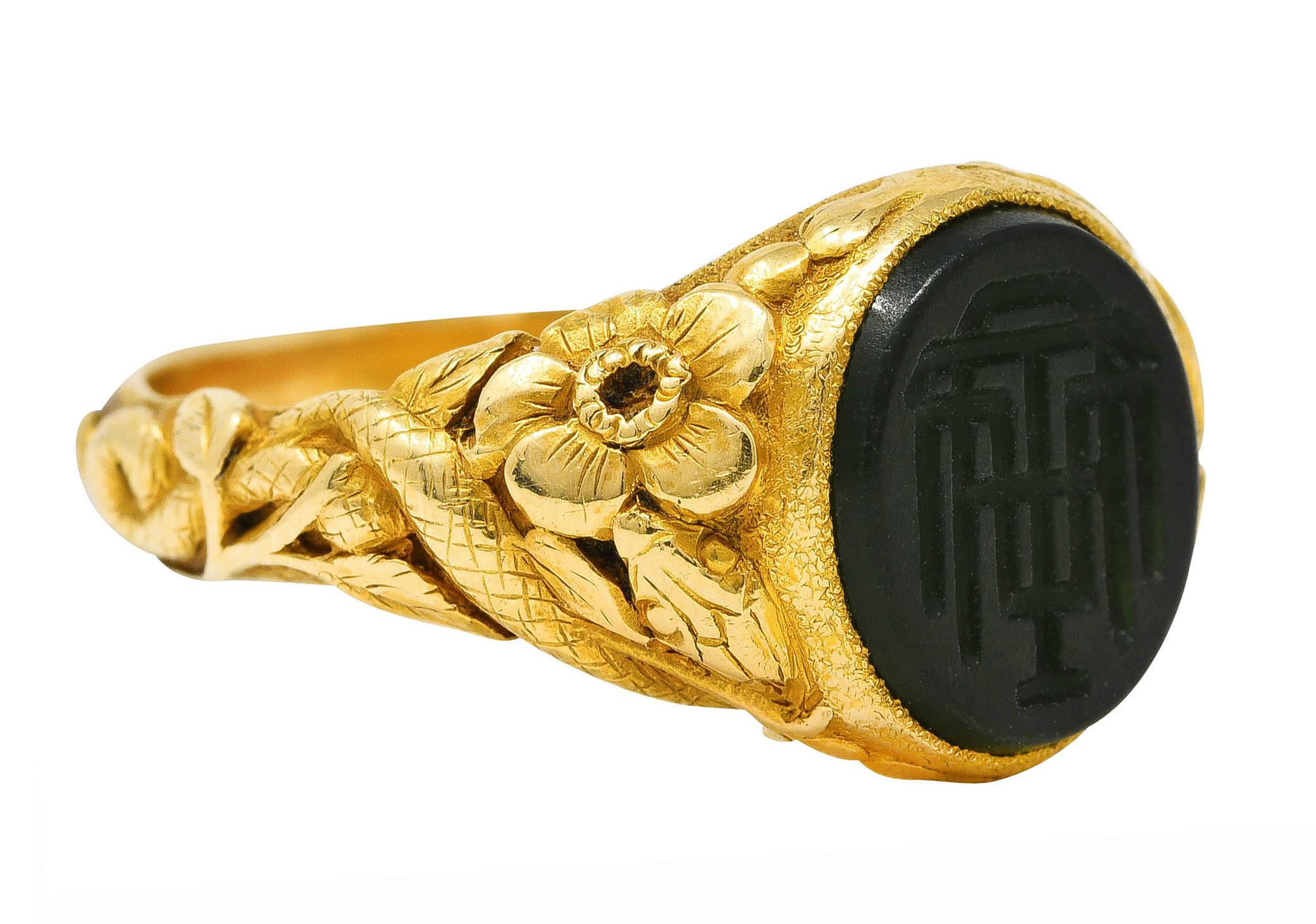 Ring centers a 8.5 x 10.0 mm nephrite jade tablet - translucent medium dark yellowish-green

Bezel set and featuring a carved intaglio of initials

Flanked by highly rendered shoulders with floral foliate and snakes

Tested as 18 karat gold

Fully