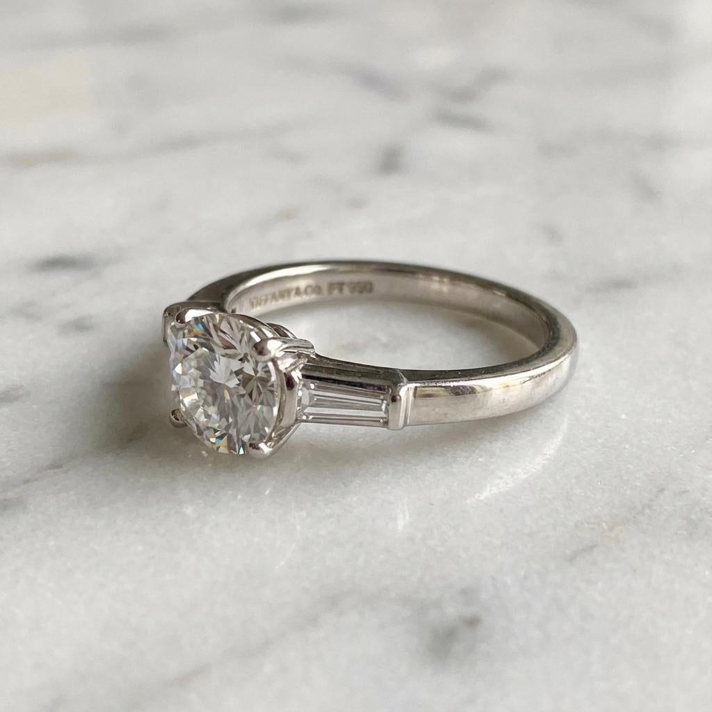 Cette exquise bague de fiançailles Tiffany & Co. met en valeur un superbe diamant rond brillant de 1,06 carat, certifié directement par Tiffany & Co. pour son authenticité et sa qualité. Le diamant central présente une pureté exceptionnelle VVS2, ce