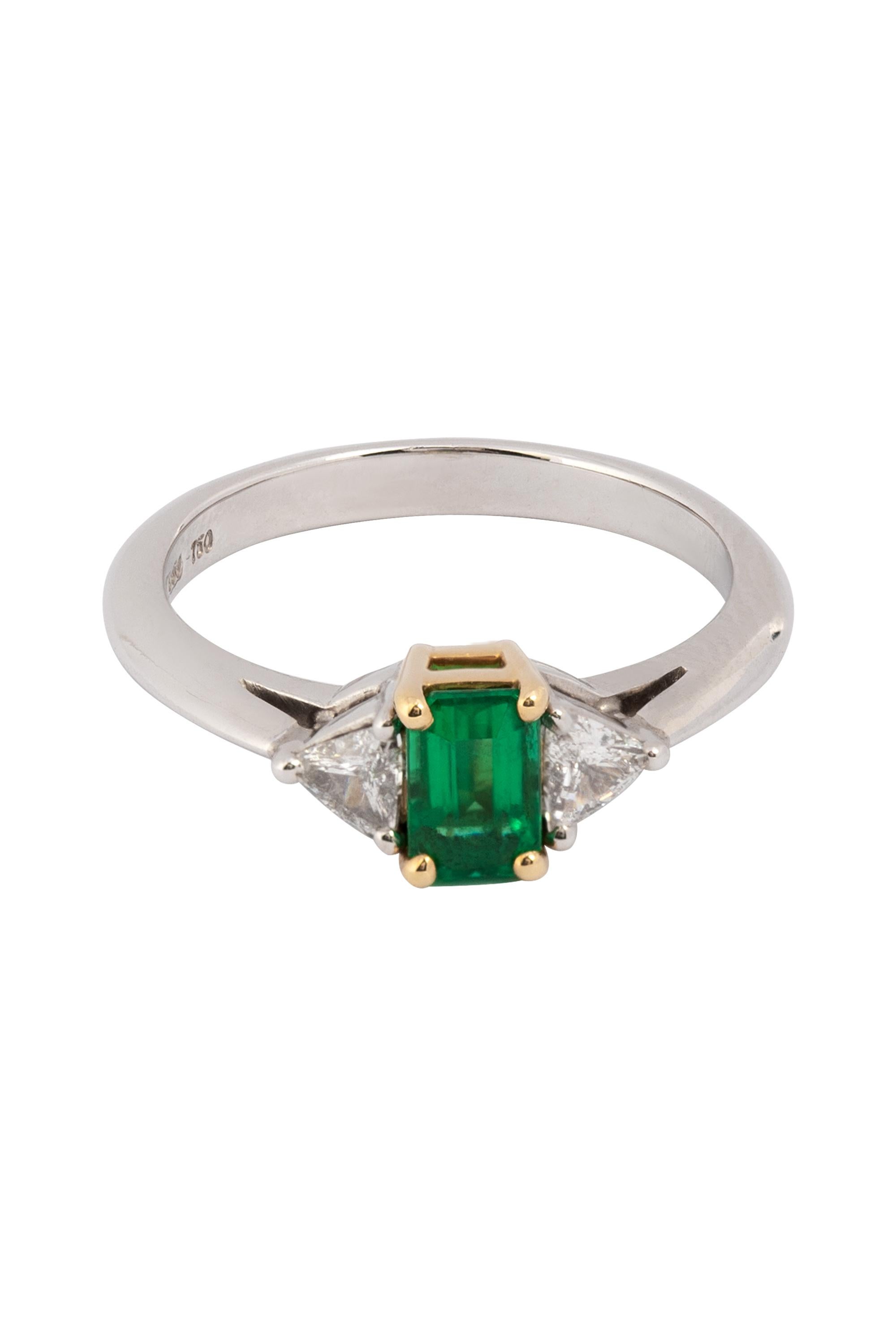 This exquisite ring from Tiffany & Co. showcases a richly saturated emerald-green gemstone, weighing approximately 0.60 carats. The vibrant emerald is elegantly complemented by two sparkling white trillion-cut diamonds, with a total diamond weight