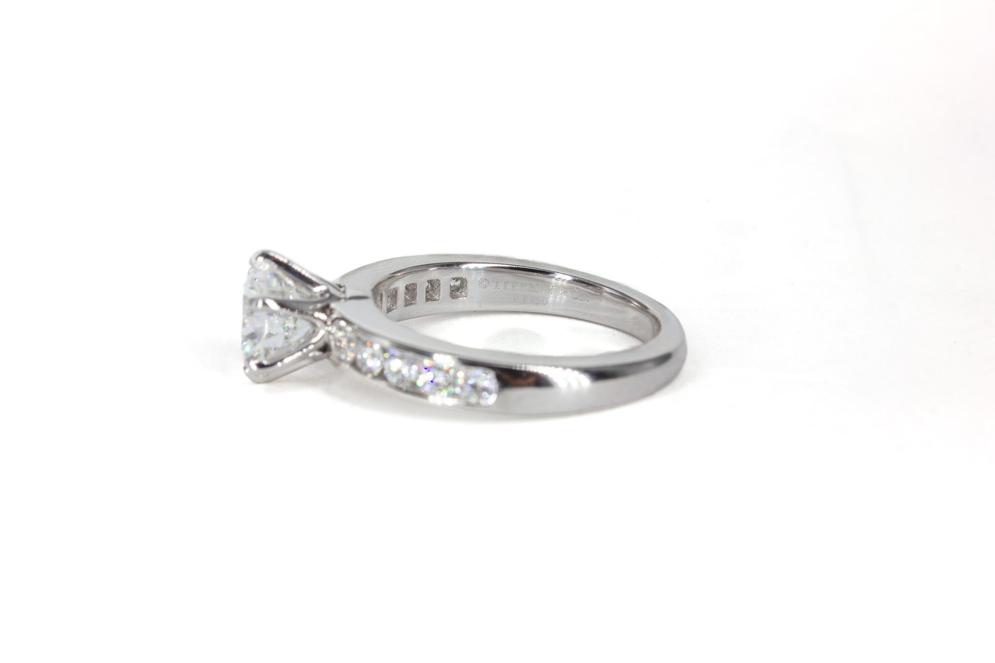 This exquisite Tiffany & Co. platinum engagement ring is a true embodiment of timeless elegance and superior craftsmanship. At its center is a dazzling 1.22 carat round brilliant diamond, certified by GIA as H color and VS1 clarity, offering