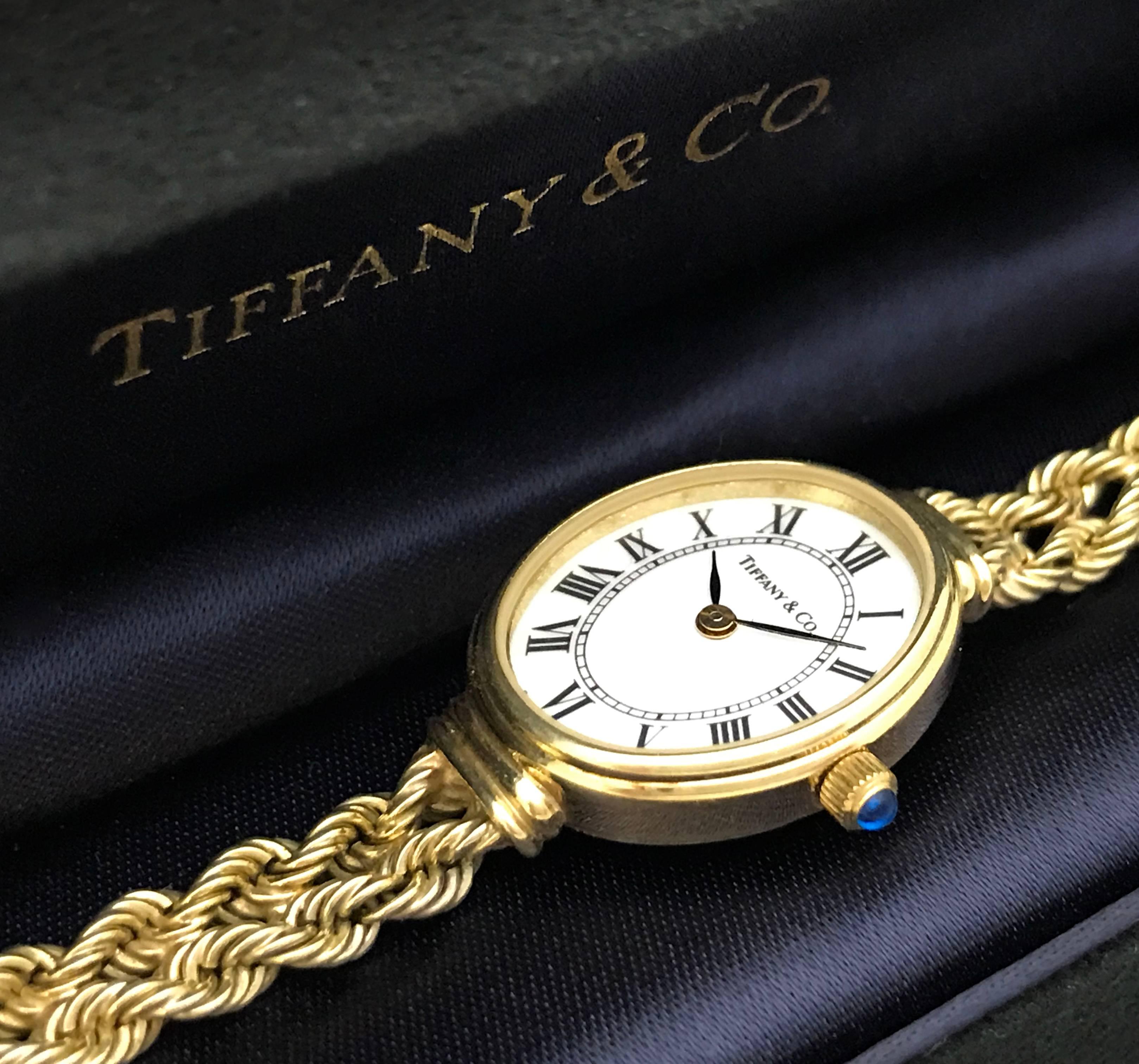 Tiffany & Co. Lady's 14k Gold Bracelet Watch, Circa 1996. The watch has an oval white dial with black enamel Roman hours, black leaf-style hour and minute hands, and a quartz movement on a double row twisted rope bracelet. Round blue sapphire detail