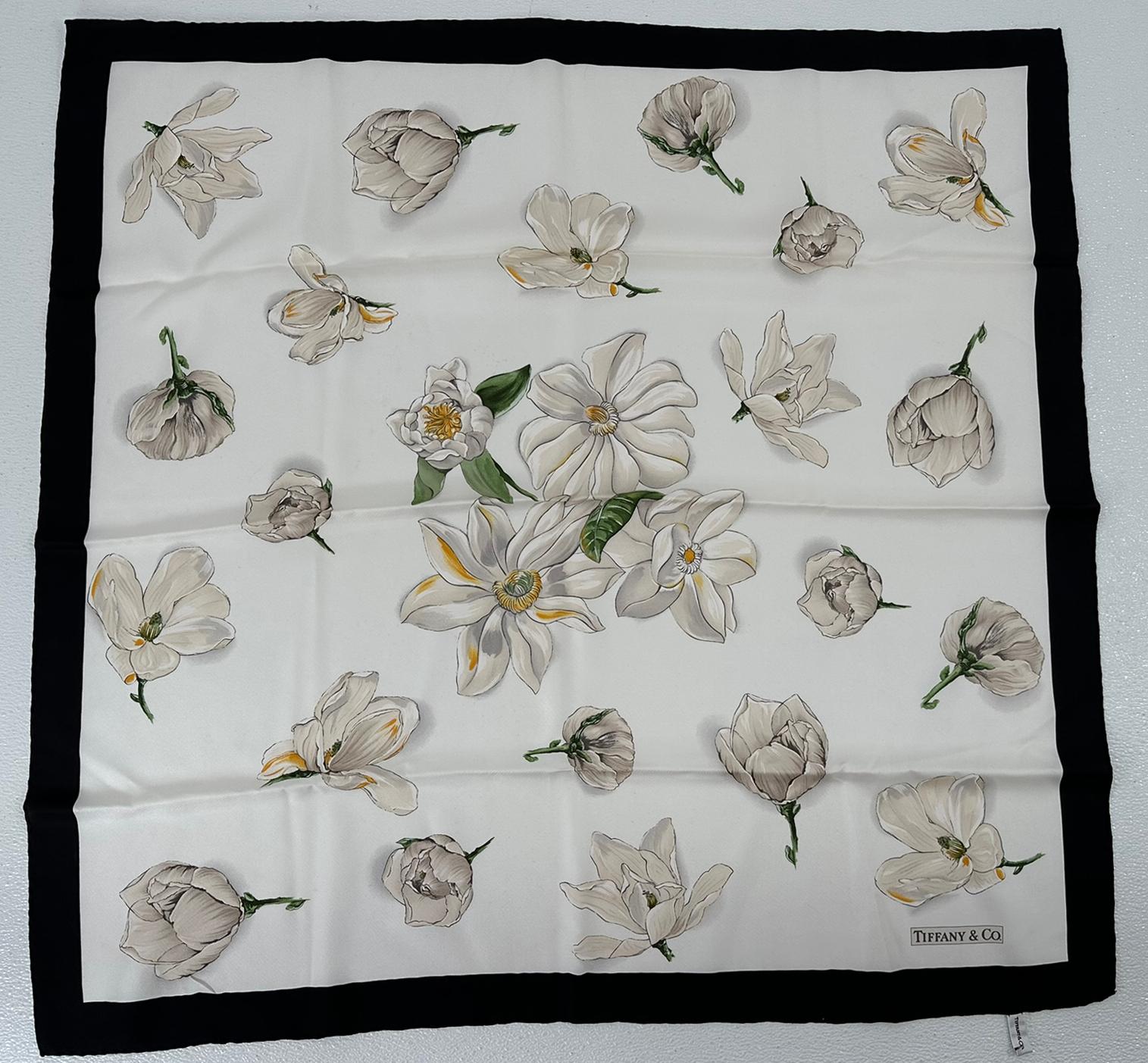 Foulard in seta Tiffany magnolia con fondo bianco e fiori di magnolia nei toni del bianco e del crema. Il bordo della sciarpa è nero, la sciarpa è firmata Tiffany & Co. Orli arrotolati a mano. Etichetta originale Tiffany attaccata. In ottime