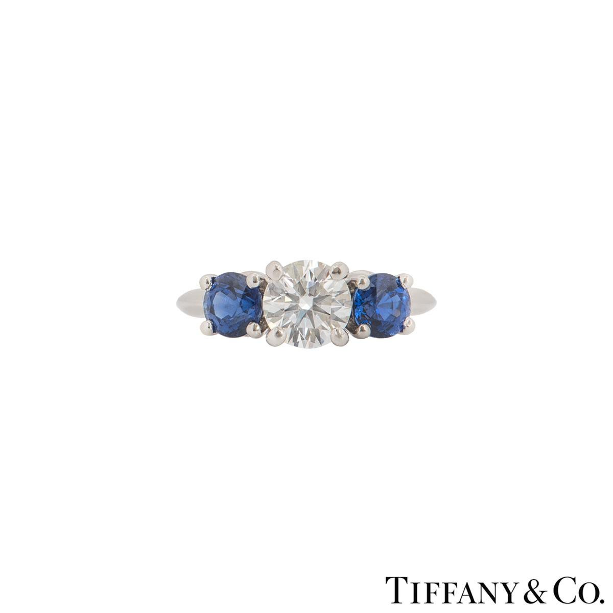A stunning platinum diamond and sapphire ring by Tiffany & Co.. The ring comprises of a round brilliant cut diamond in the centre with a sapphire stone on each side all in a four claw setting. The round brilliant cut diamond has a weight of 1.06ct,