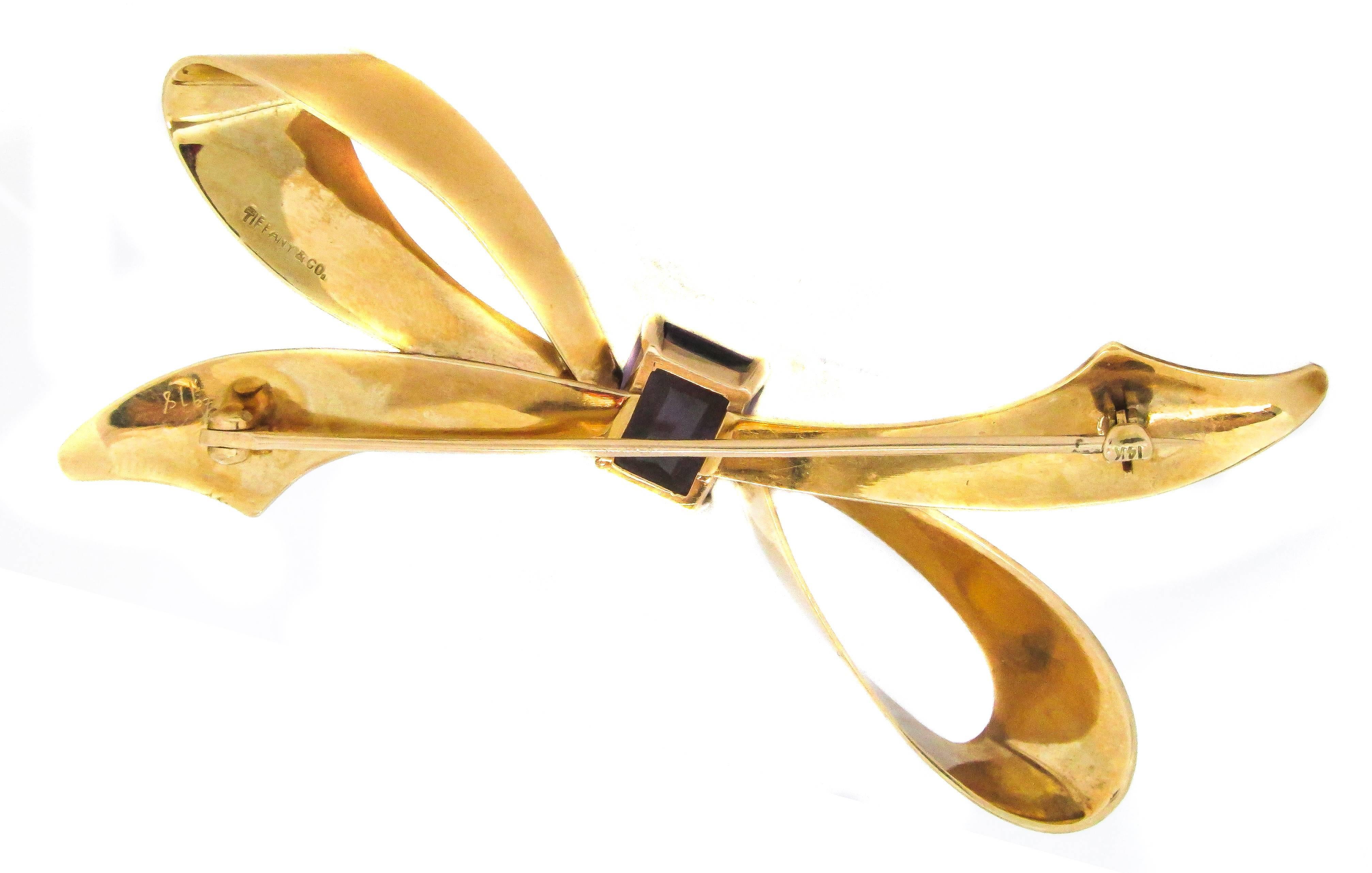Retro 14 karat yellow gold and amethyst bow brooch by Tiffany & Co. An emerald cut Amethyst is centrally set in high polished gold giving this piece of jewelry a classy and stylish look. The light weight of this beautiful brooch makes it easy to