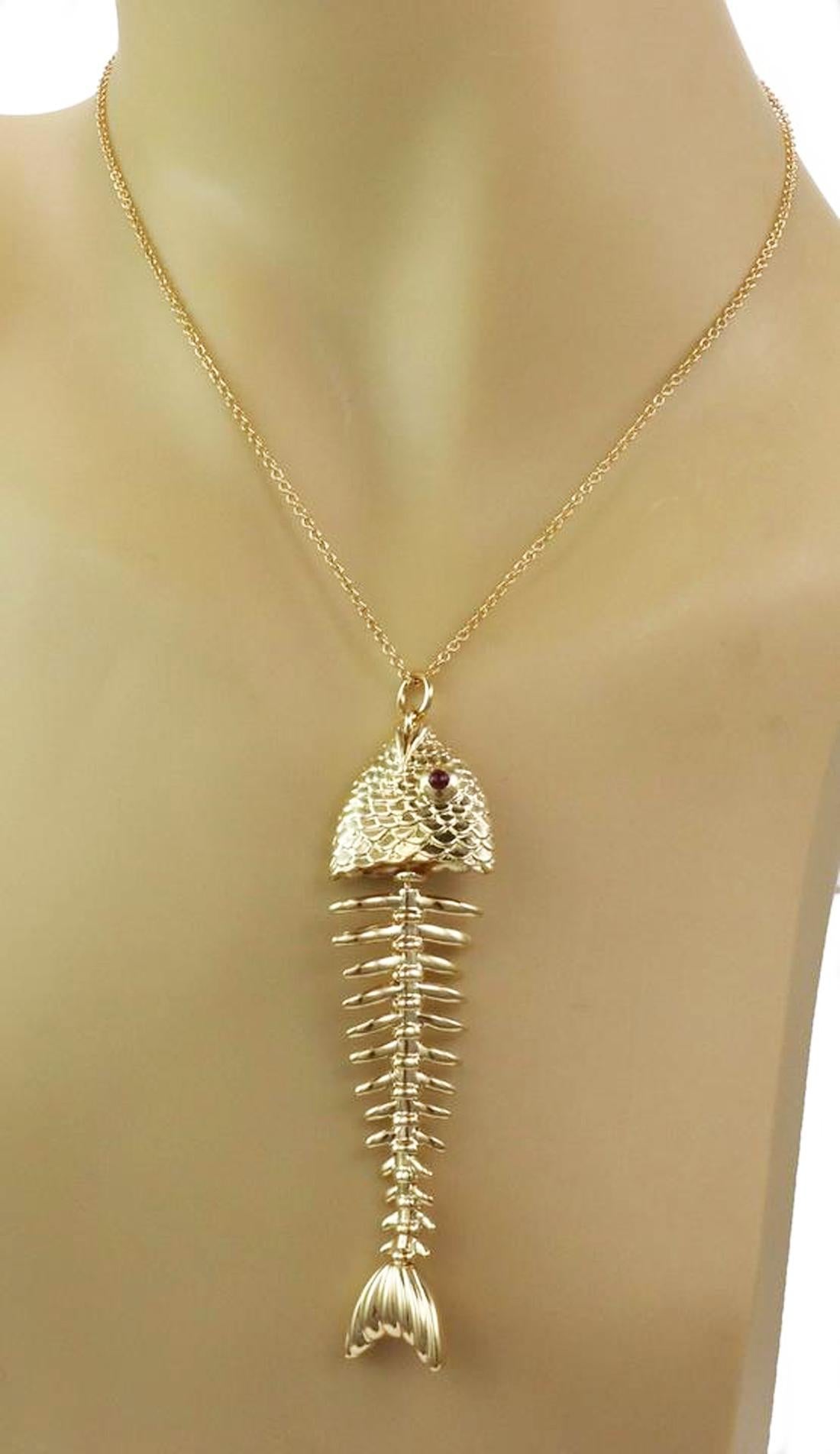 This rare authentic pendant and chain from Tiffany & Co. It is crafted from 18k yellow gold with a polished finish. The pendant features a large fish deboned with its scaled head attached to the bone and has cabochon ruby eyes. the tail is also