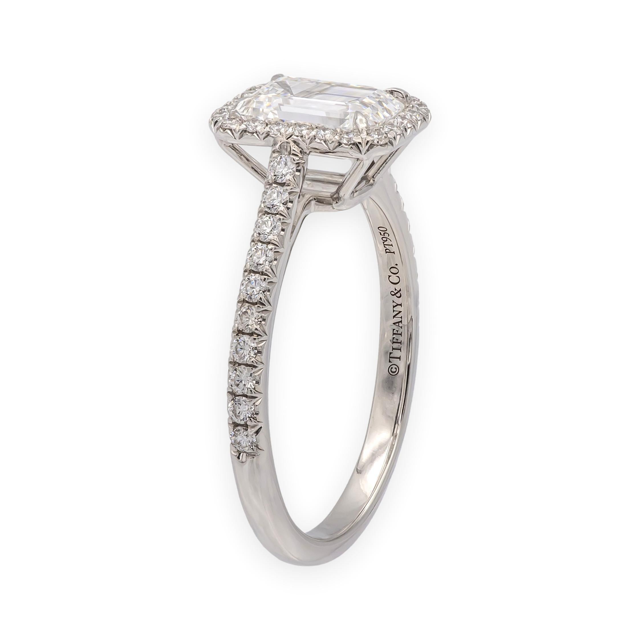 This Tiffany & Co. Soleste engagement ring features a 1.12-carat emerald-cut diamond at the center, graded F color and VVS2 clarity. The center stone is surrounded by a halo of round brilliant diamonds and additional pavé-set diamonds along the