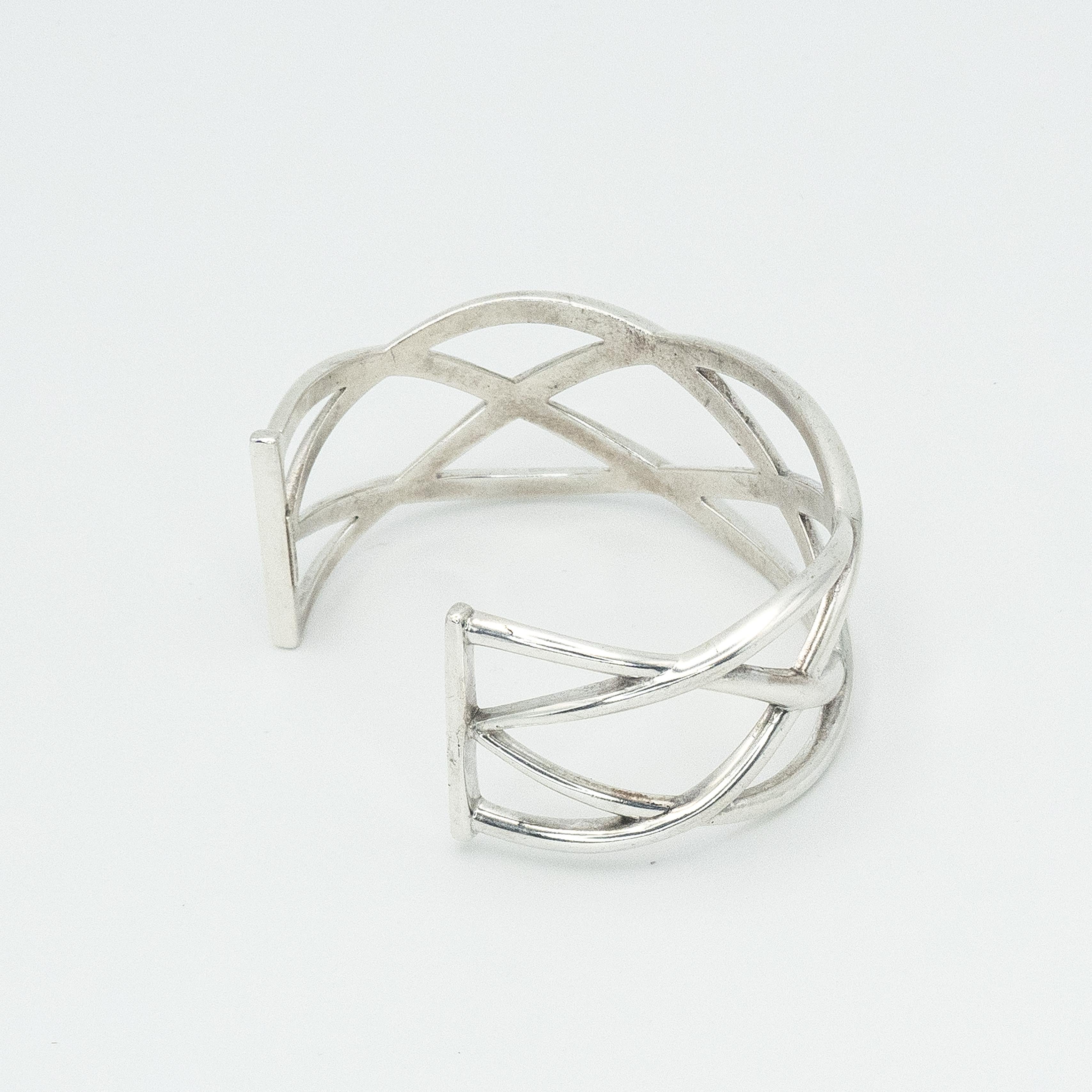 An elegant and iconic design by Tiffany & Co., this sterling silver cuff features a bold openwork motif of overlapping criss-cross lines, creating a striking lattice effect. The polished finish enhances its sculptural form, reflecting light with