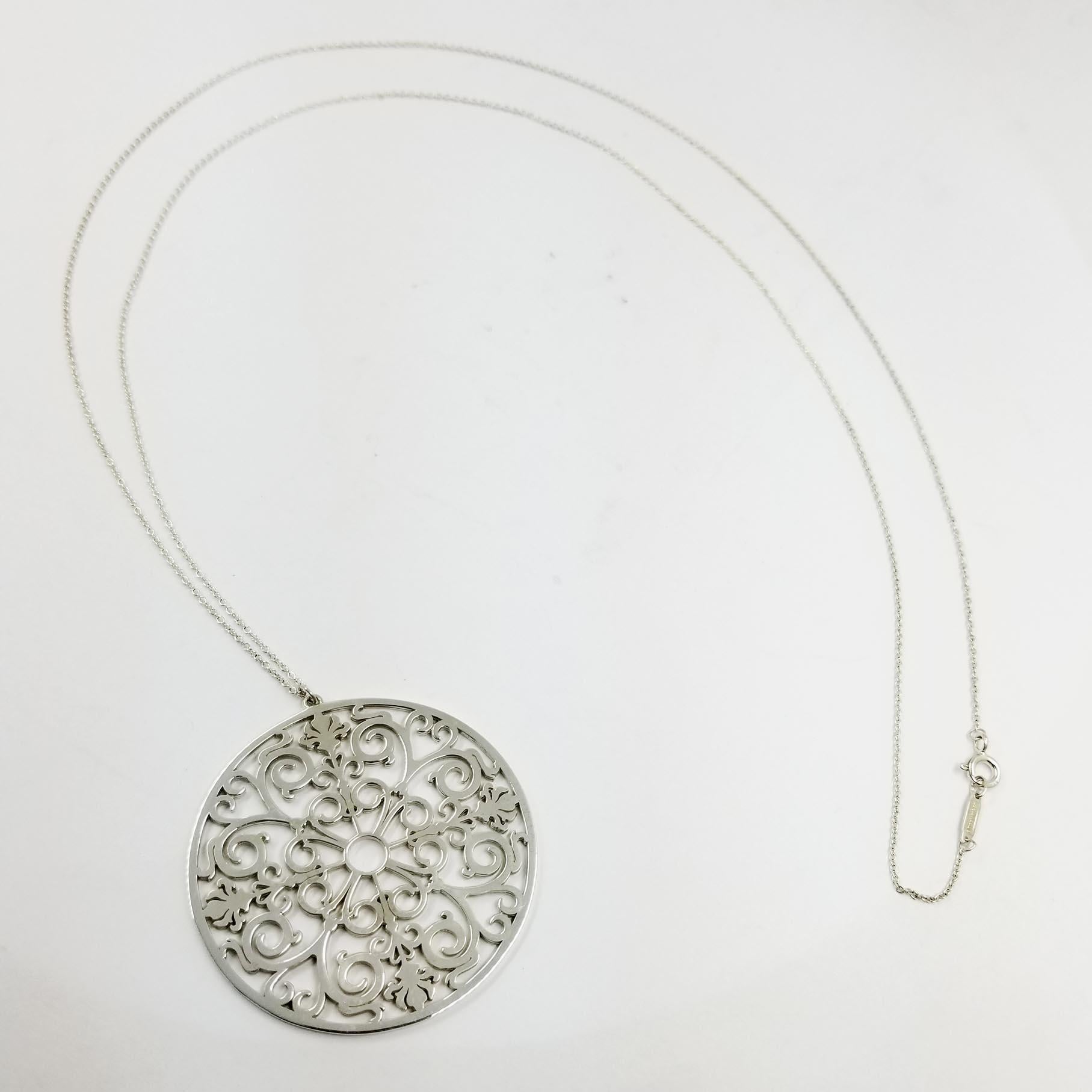 This sterling silver medallion necklace is designed by Tiffany & Co. The thin 30 inch chain is marked AG925 TIFFANY&Co. The pendant is designed with open scroll work, and measures approximately 2 inches in diameter. It has been professionally