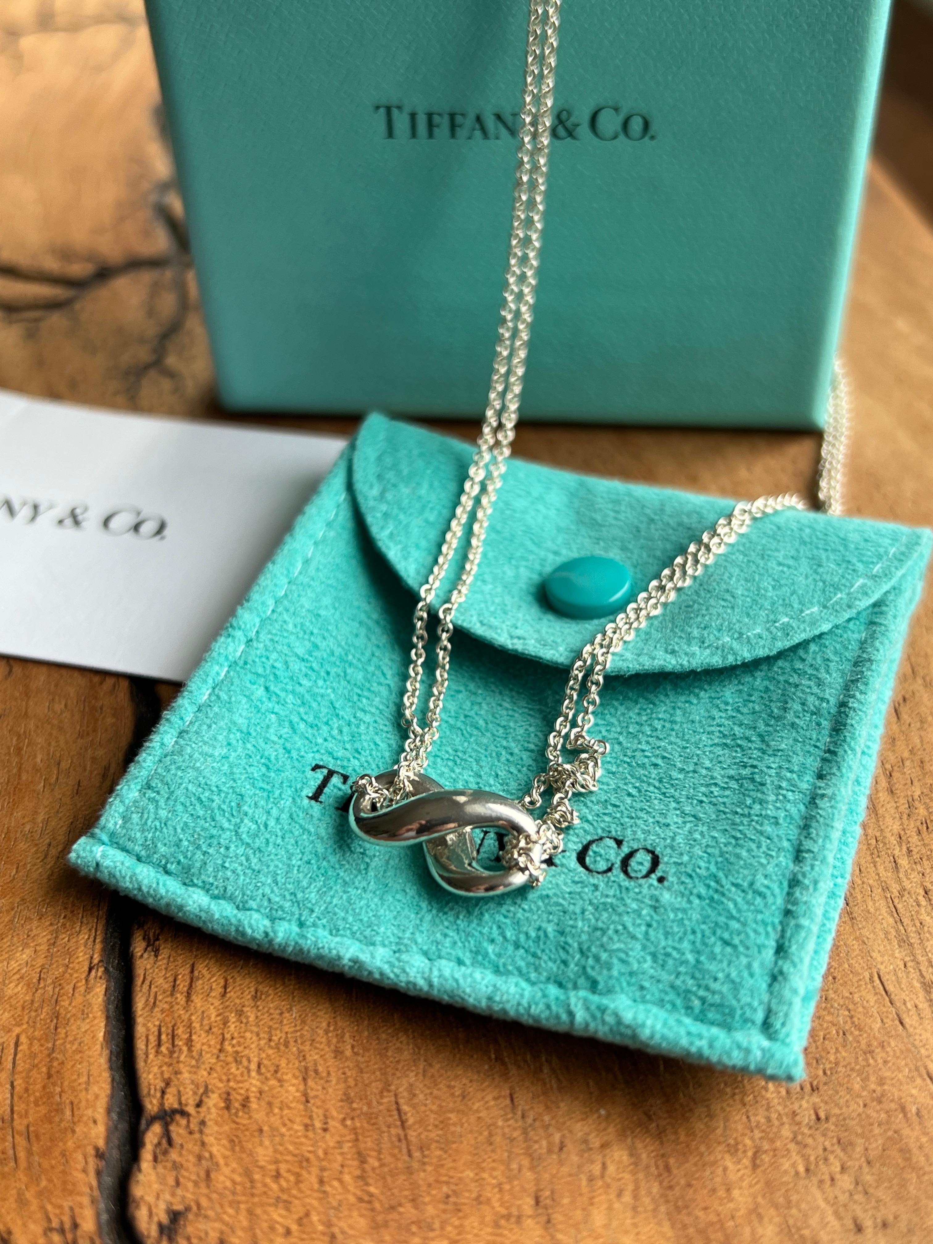 This contemporary Tiffany & Co. necklace features the iconic infinity symbol, crafted in sterling silver and suspended from a delicate double-strand 2mm cable chain with spring ring clasp. The charm measures approximately 3/4 by 3/8 inches and is