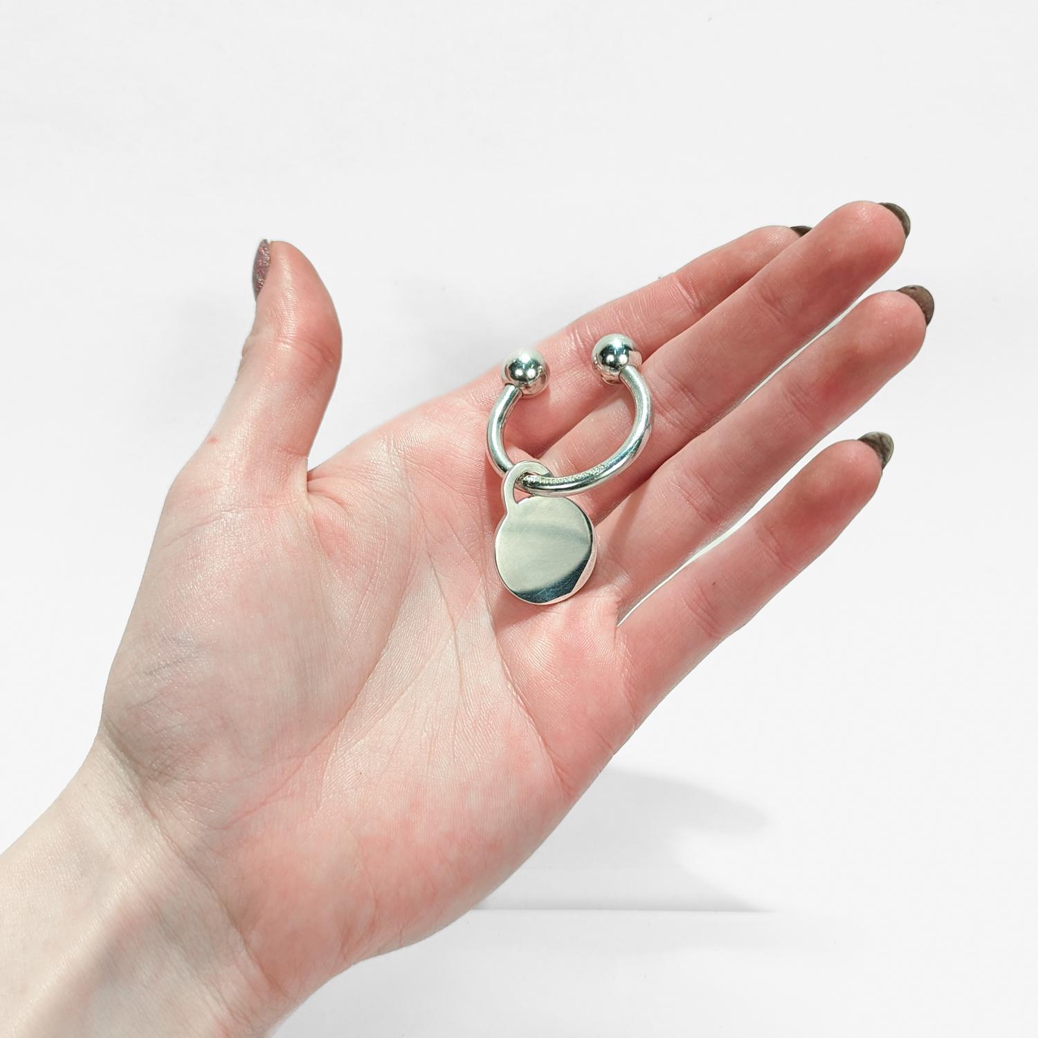 Un ottimo portachiavi.

Da Tiffany & Co.

In argento sterling.

Composto da una barra circolare con terminali a forma di palla e una targhetta rotonda allegata.

Uno dei terminali a forma di sfera è filettato e può essere aperto per aggiungere o