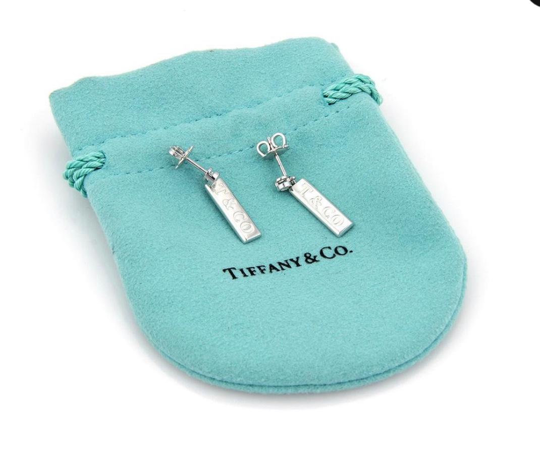 A true classic authentic pair of earrings by Tiffany & Co.
These authentic Tiffany & Co earrings are finely crafted from solid 18k white gold. They feature dangling long bars engraved with the iconic logo T&Co. along with 2 sparkling diamonds above