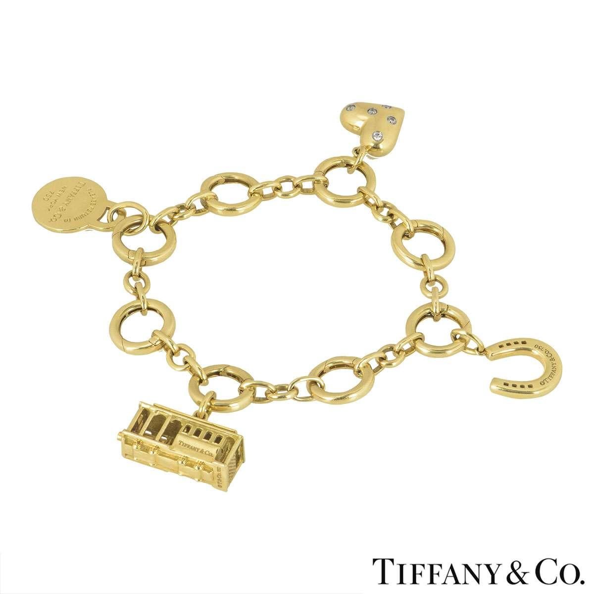 An 18k yellow gold charm bracelet by Tiffany & Co. The bracelet has 4 charms which consist of a diamond heart, a horseshoe, a tram and a Return to Tiffany & Co motif. The bracelet is made up of circular links, each featuring a push spring clasp