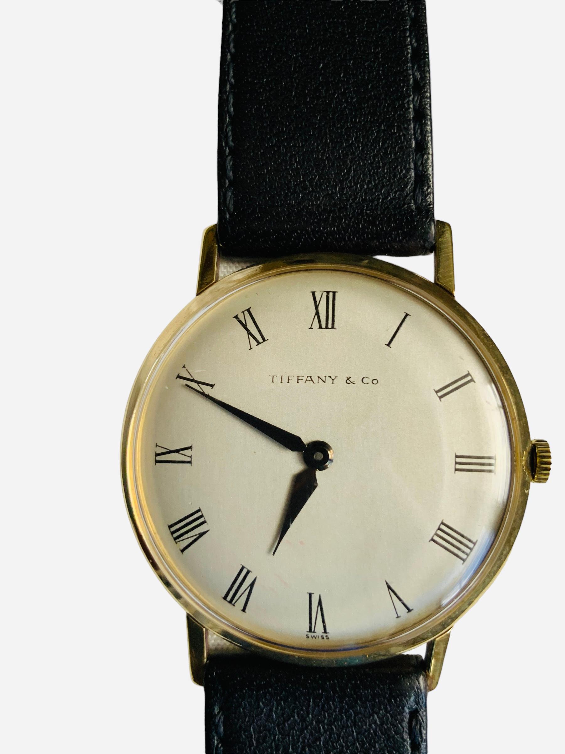 Questo è un orologio da polso da uomo Tiffany & Company. Presenta una cassa rotonda in oro 18 carati e un quadrante grigio biancastro con numeri romani e lancette delle ore e dei minuti di colore nero. Sotto le ore 6, il marchio è svizzero. Un vetro