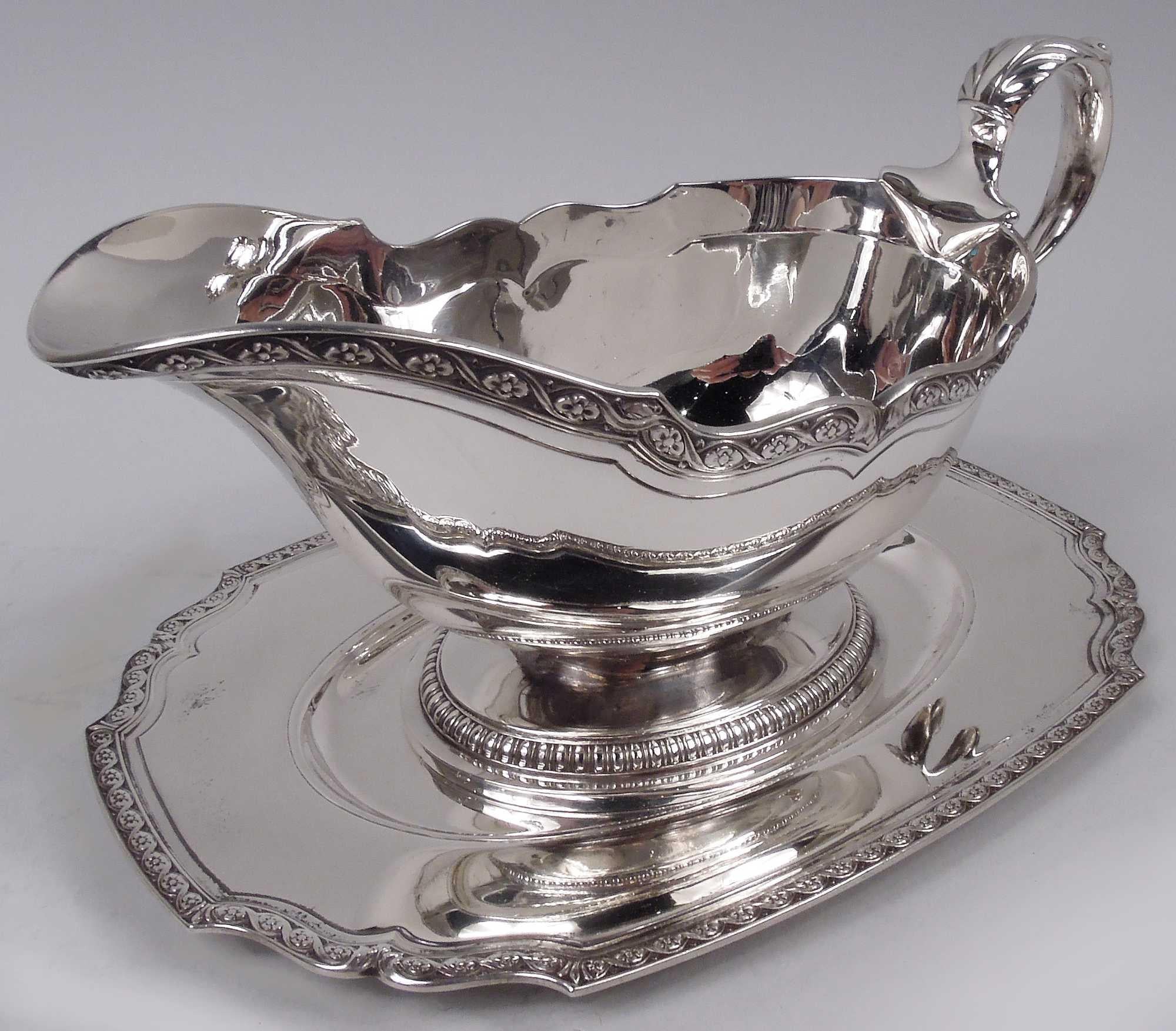 Edwardian Classical sterling silver gravy boat on stand. Made by Tiffany & Co. in New York, ca 1924. Boat: Ovoid bowl with scrolled helmet mouth, and leaf-capped high-looping double-scroll handle; raised foot. Stand: Rectilinear with scrolled
