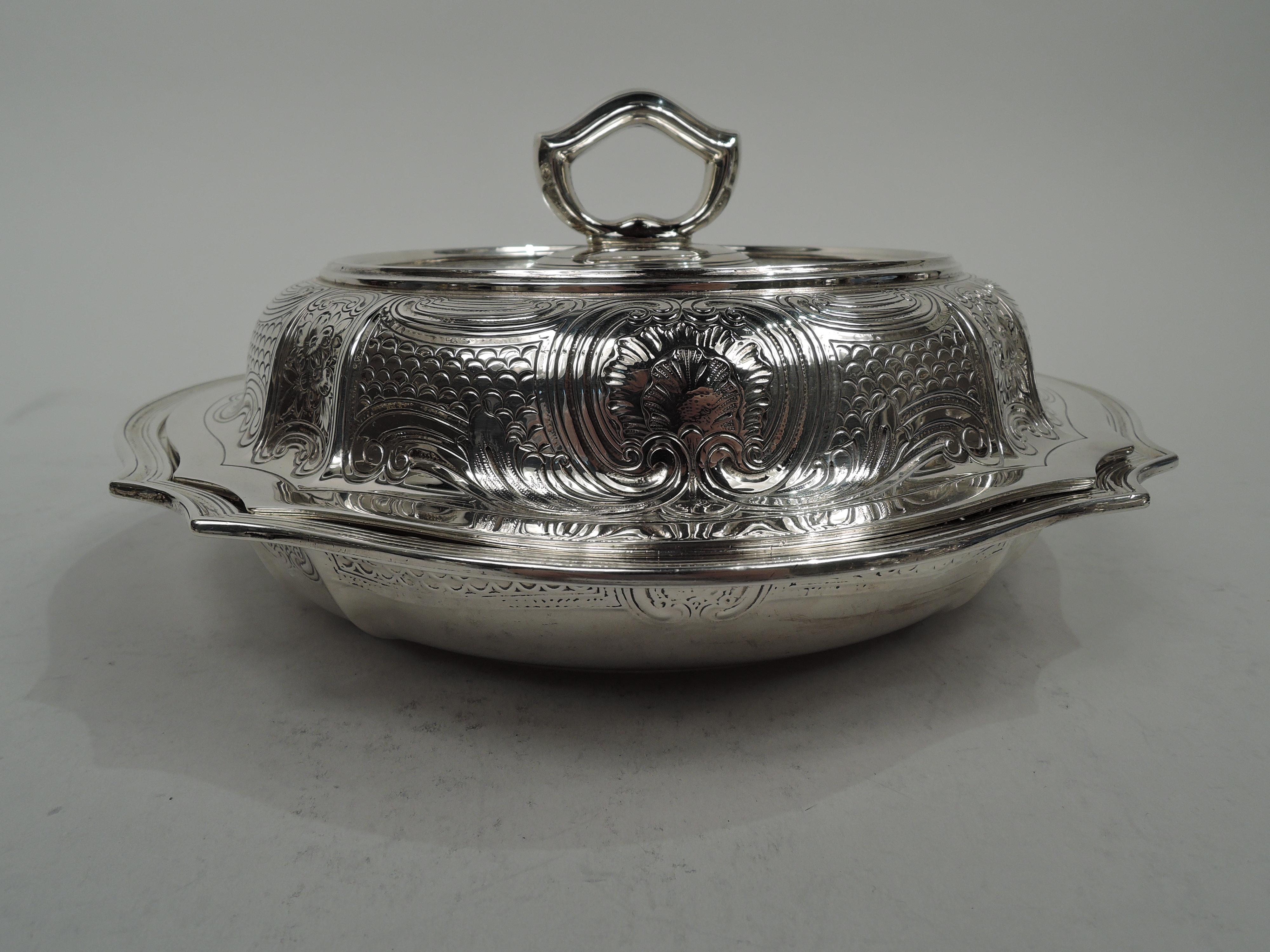 Edwardian Modern classical sterling silver serving bowl. Made by Tiffany & Co. in New York, ca 1910. Round and serpentine with tapering sides and reeded rim. Cover raised with tapering sides and plain and flat rim. Dense engraved scrolls, flowers,