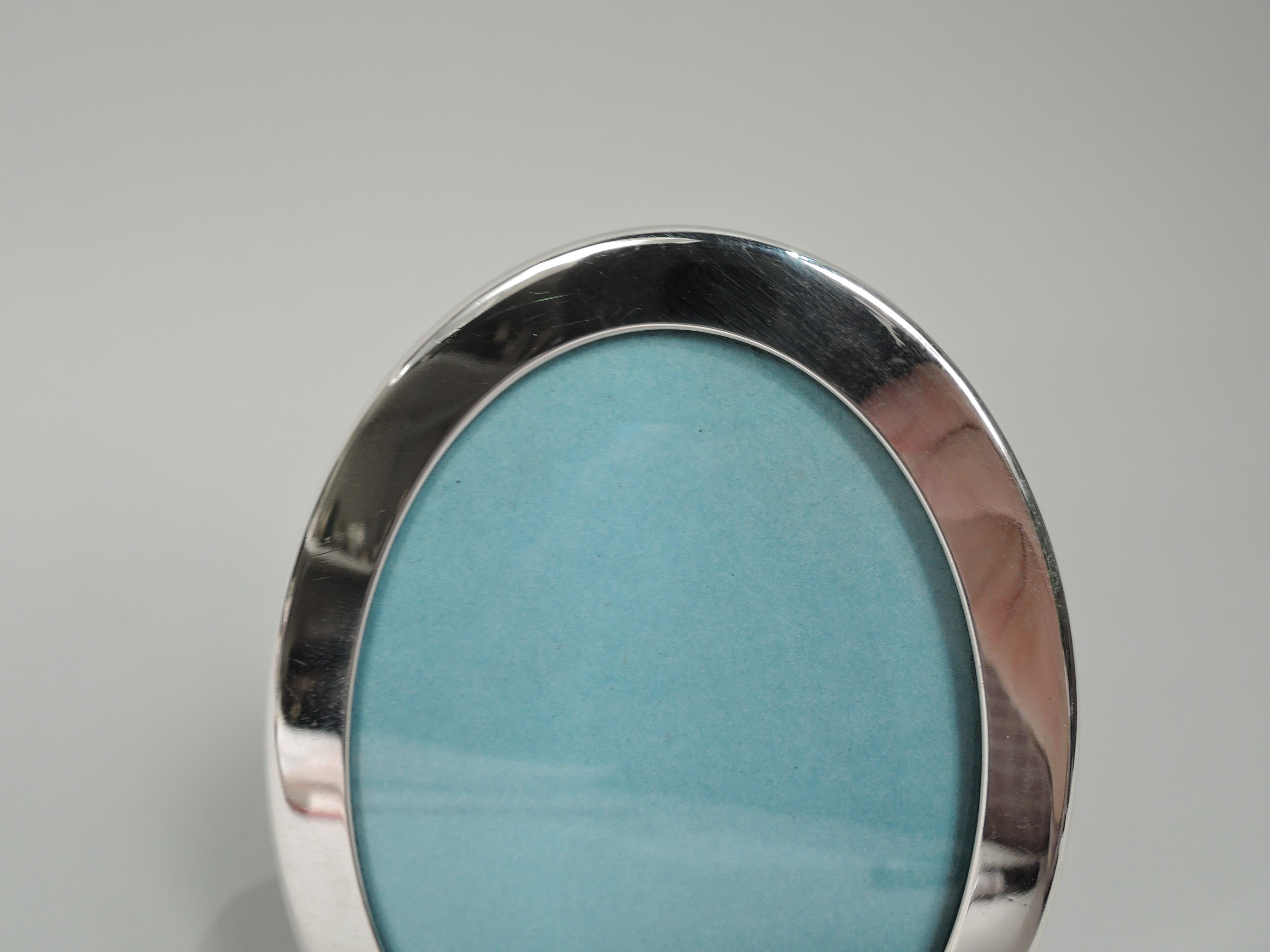 Small sterling silver picture frame. Retailed by Tiffany & Co. in New York. Oval window and flat surround. With glass, eggshell-blue paper liner, and laminate back and hinged easel support. Marked “Tiffany & Co. / Sterling” and post-1967 Italian