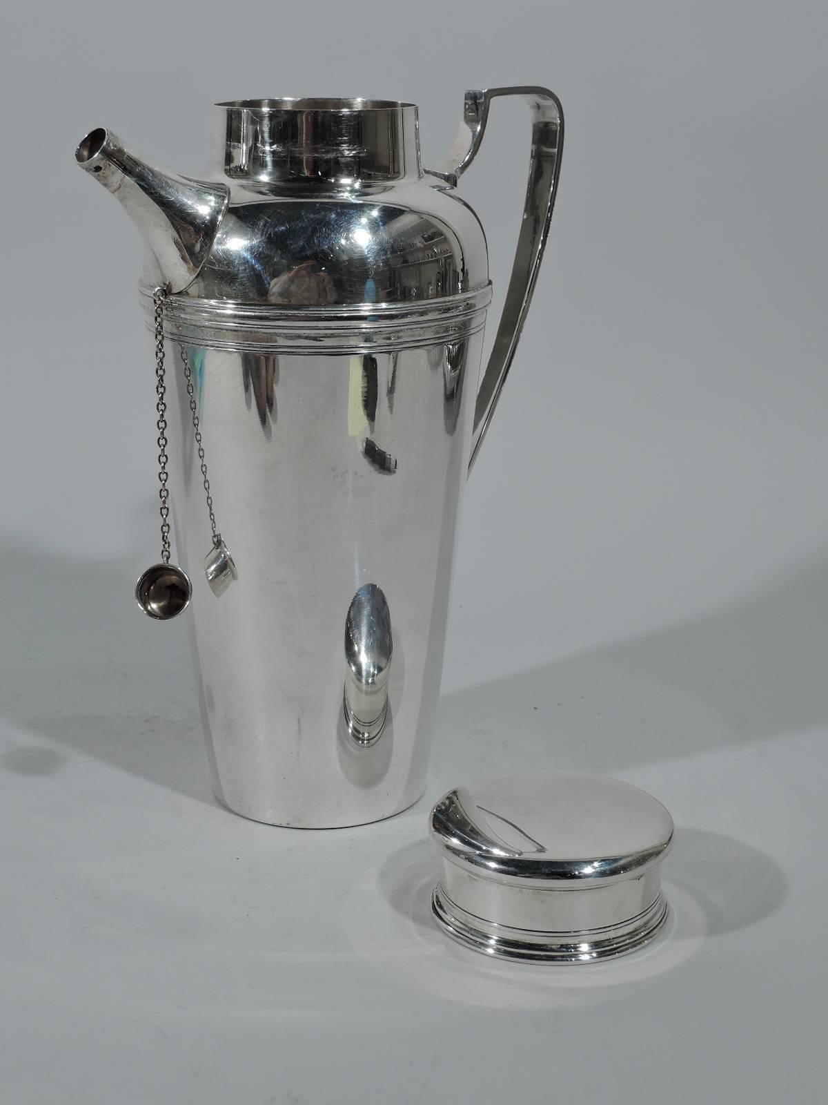 Modern sterling silver cocktail shaker. Made by Tiffany & Co. in New York. Tapering sides, short neck, flat and overhanging cover, scroll bracket handle, and diagonal spout with built-in strainer and chained cap. Spare with incised bands. Hallmark