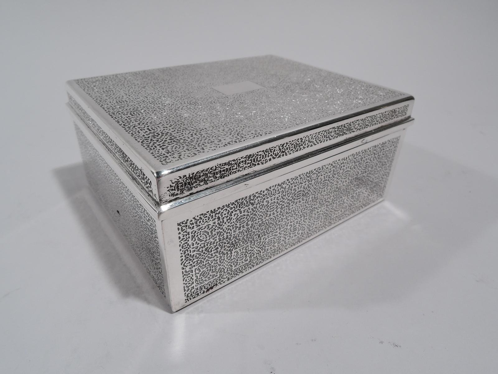 Small modern sterling silver box. Made by Tiffany & Co. in New York. Rectangular with straight sides and angular corners. Cover flat and hinged. Fine and dense acid-etched all-over scrollwork mesh; cover top has rectangular cartouche (vacant). Box