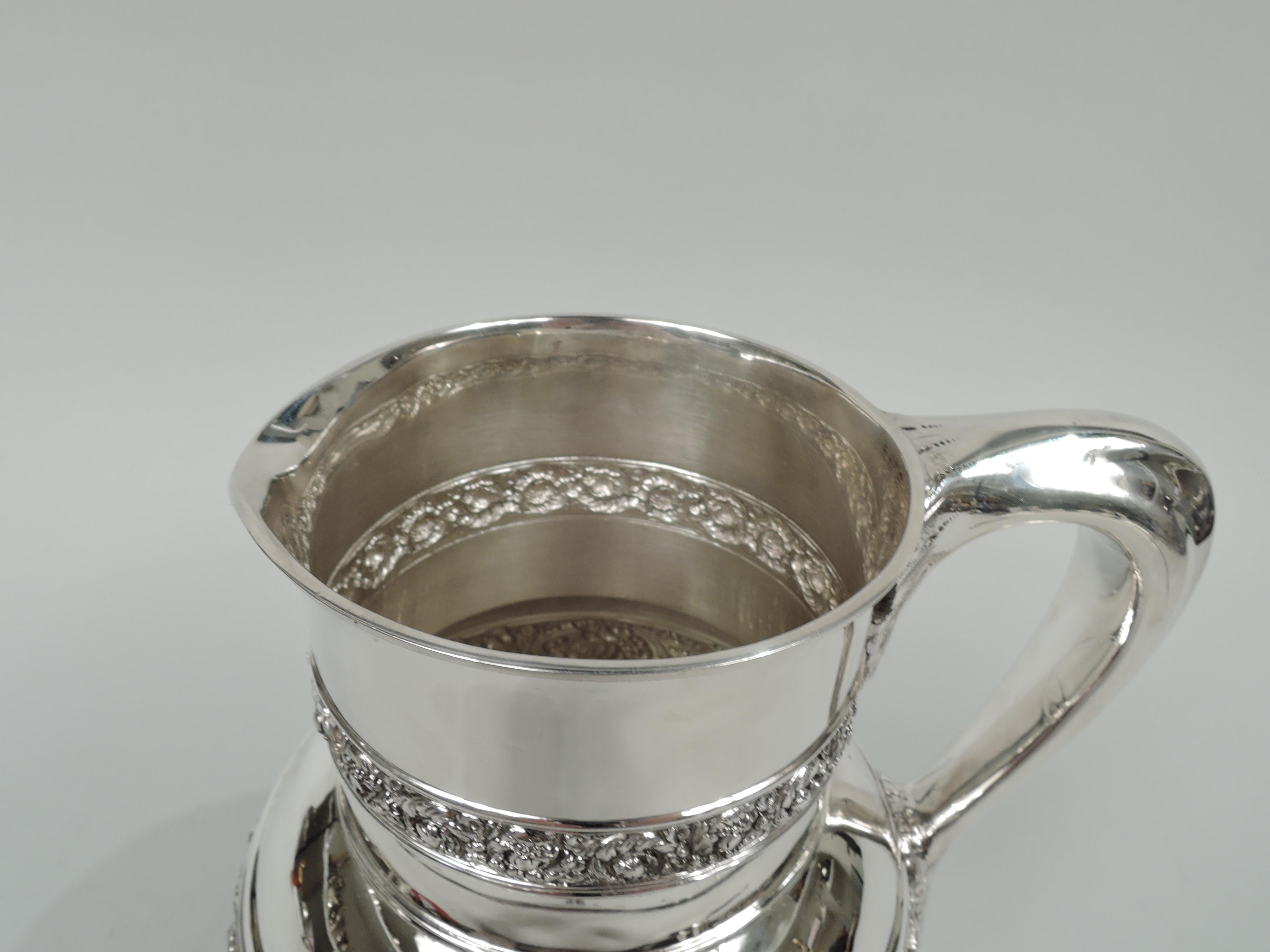 Olympian sterling silver water pitcher. Made by Tiffany & Co. in New York. Globular with drum-form neck, small lip spout, and leaf-mounted scroll bracket handle. Exterior encircled with chased and embossed frieze of love-making and harp-strumming