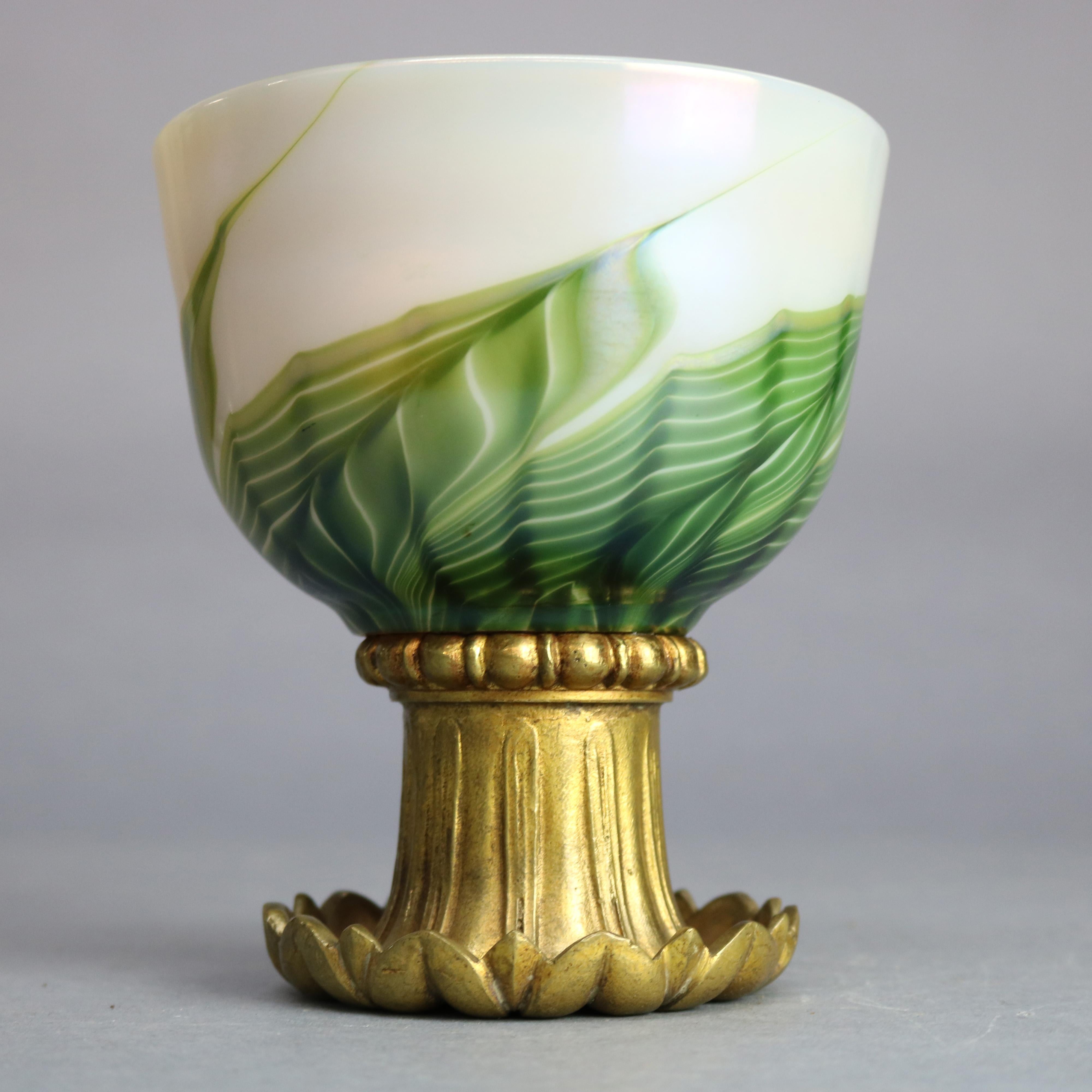 An an antique Art Nouveau ceremonial chalice by Tiffany Studio offers Favrile art glass bowl having green, gold and white pulled feather design and gold wash interior, raised on gilt bronze foliate form base, maker signed and ambigram as