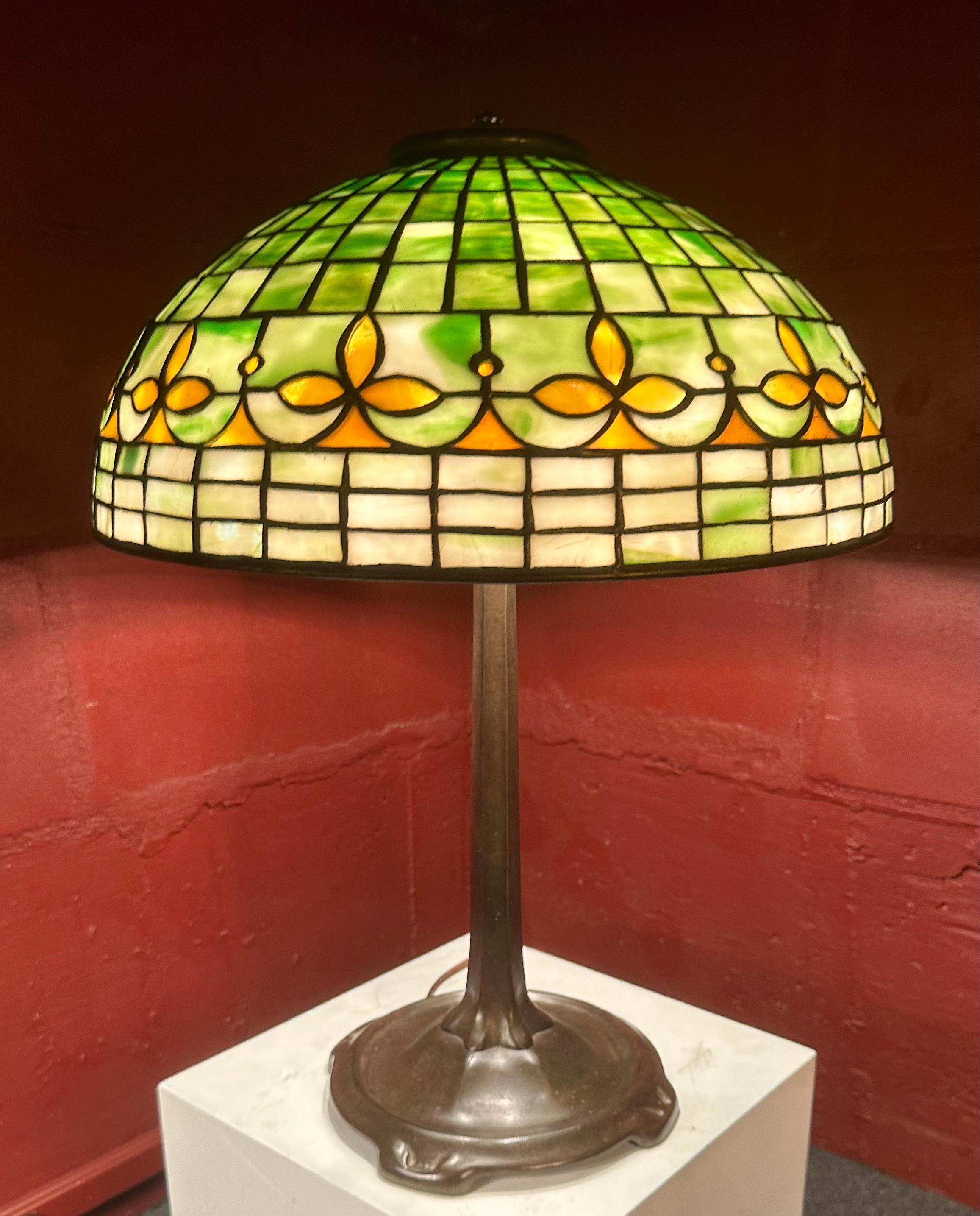 A 16” Fleur de Lis geometric lamp shade signed with the early “tag” reading “Tiffany Studios, New York”.The shade rests on the Art Nouveau stick base signed “Tiffany Studios, New York”.The lamp is complete with heat cap. It is newly rewired & works
