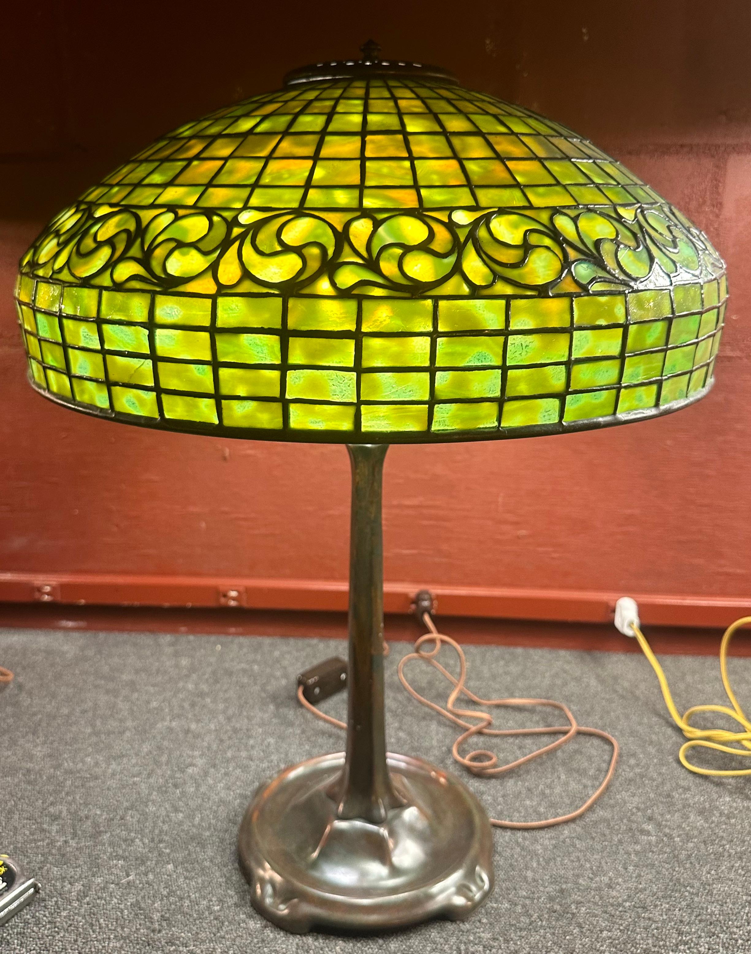 An original Tiffany Studios 18” diameter geometric table lamp with a circumferential band of “whirling leaves” at its shoulder. It is signed with a small tag that reads “Tiffany Studios, New York”.The shade rests on a Tiffany Studios “stick” base