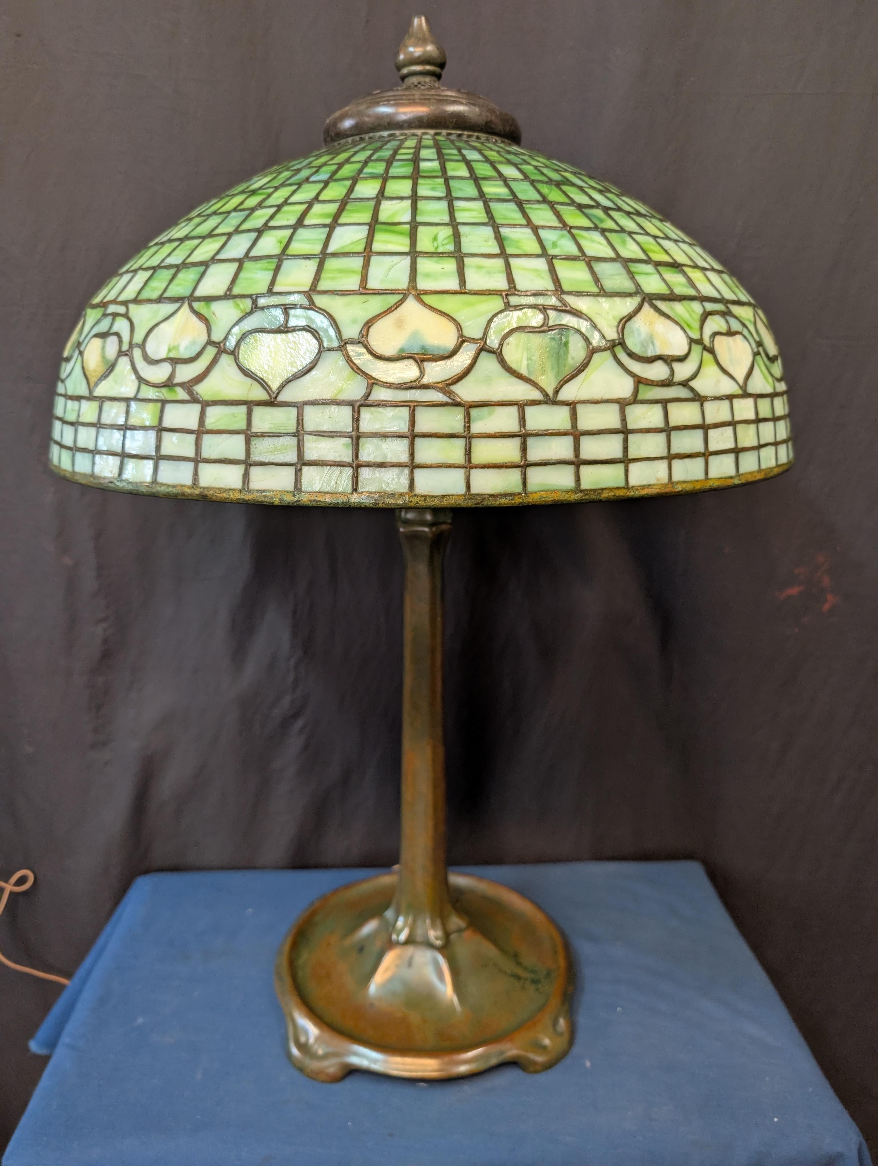 This vintage genuine Tiffany Studios table lamp is circa the early 1900’s. Featured is a beautiful tranditionally designed shade with a geometric pattern accented by a acorn motif. Shade displays a wonderful veriety of green colors that range from