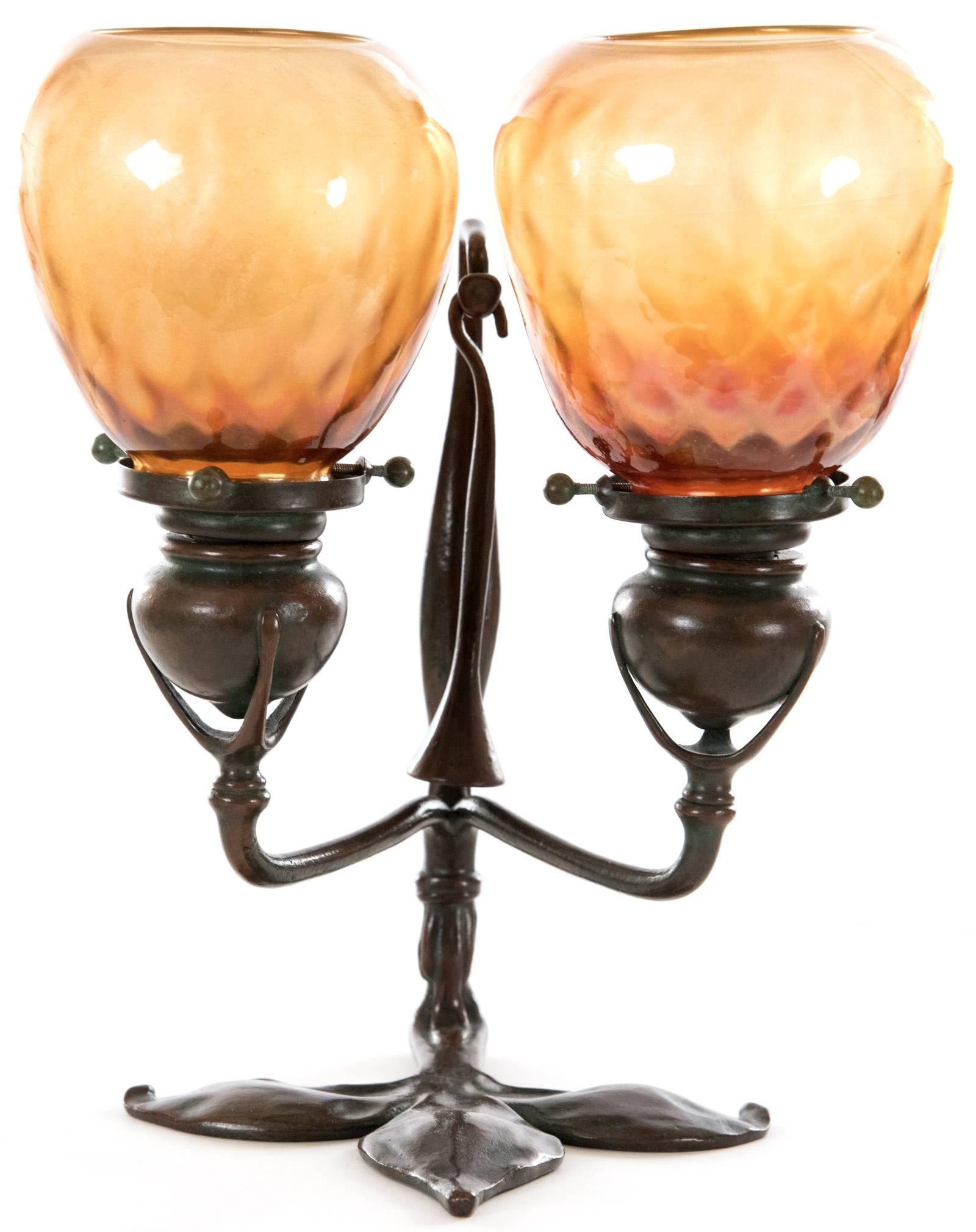 An early 20th century, circa 1910, Tiffany Studios bronze and favrile glass two-light table lamp with a fleur-de-lis formed base, including the original suspended bronze candle snuffer. The original candle stick sconces are fitted with favrile glass