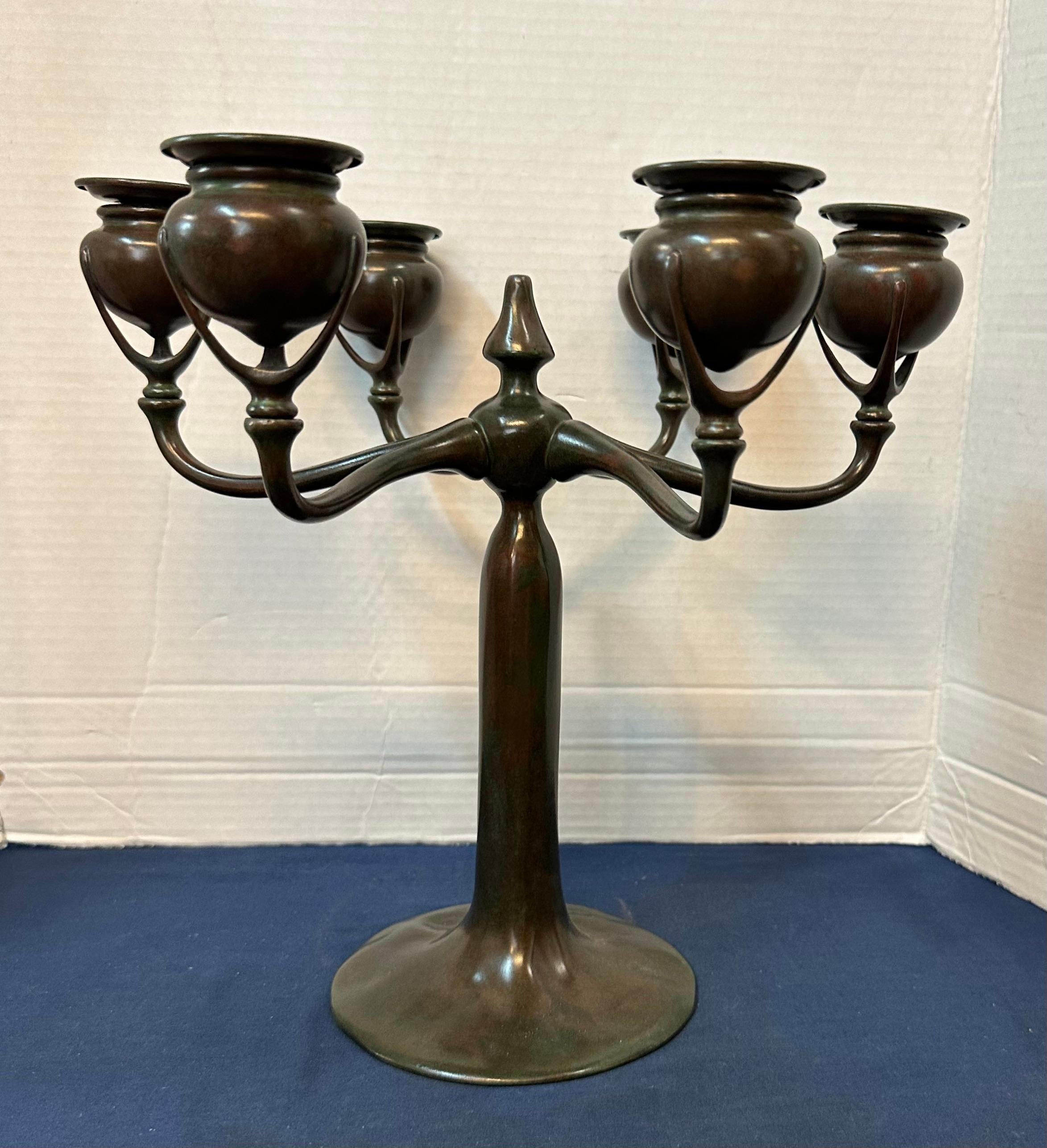 Questo autentico candelabro vintage in bronzo di Tiffany Studios risale ai primi anni Venti. Presenta sei portacandele a forma di urna bulbosa che decorano e accentuano ogni braccio esteso.
La patina originale color cioccolato ne evidenzia l'aspetto