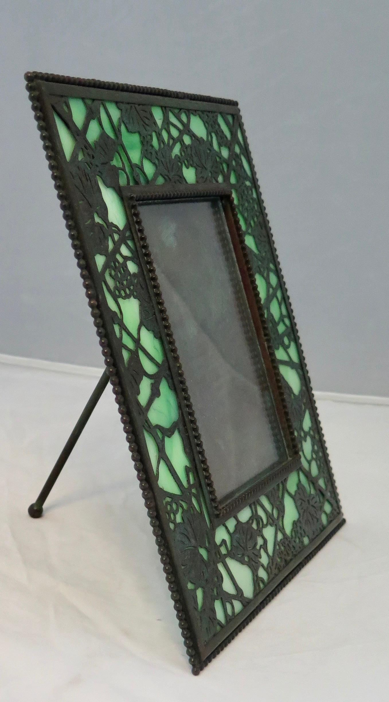 This vintage Tiffany Studios, New York photo frame is designed in the grapevine motif & dates from the early 20th century. The patinated bronze easel backed frame features an open work grapevine bronze overlaying Tiffany green favrile art glass.