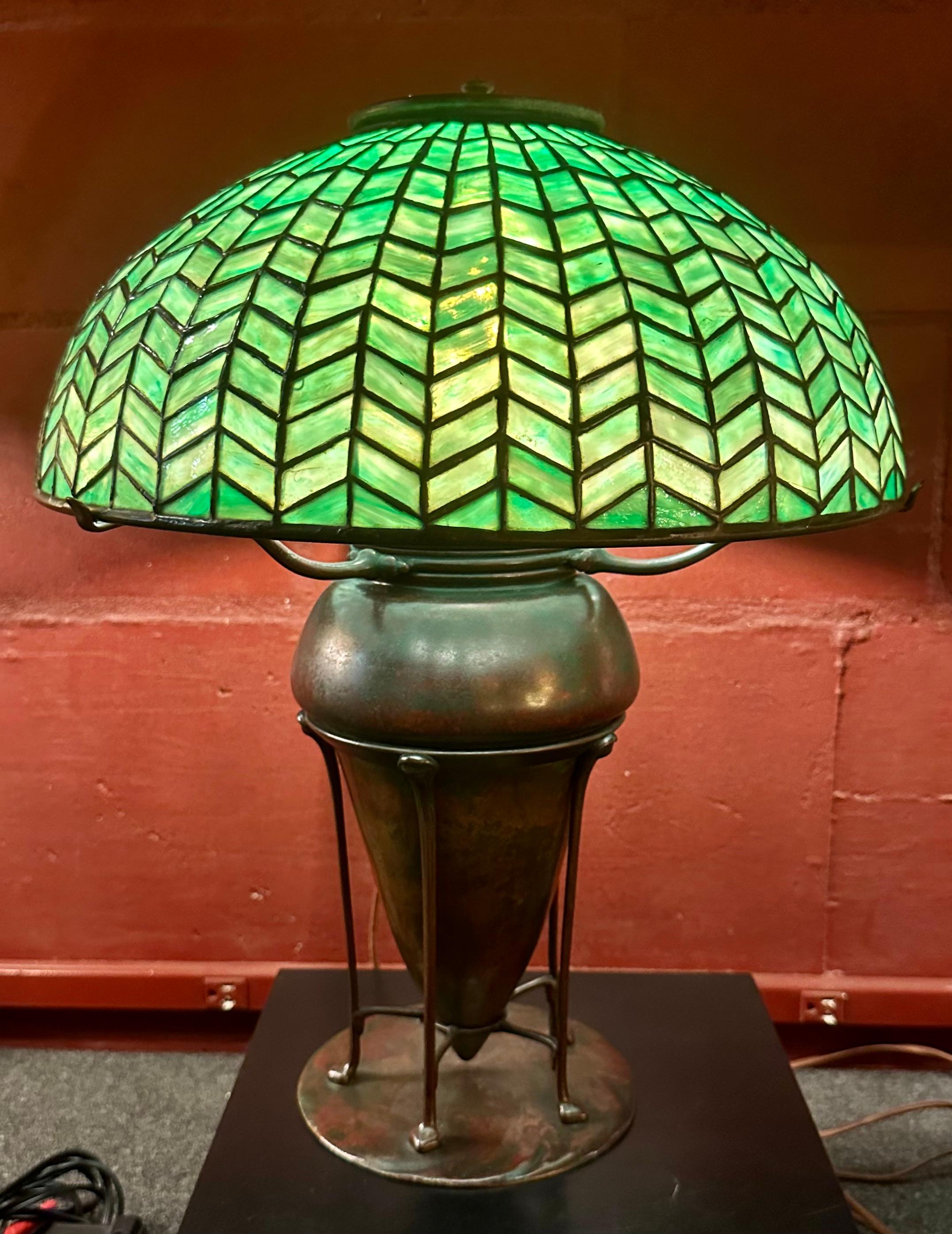 This Tiffany Studios lamp shade is referred to as a Herringbone or Zipper shade.It has a 16” diameter & is signed “Tiffany Studios, New York on an early tag. The shade rests on a spider on an early kerosene base with an original conversion to