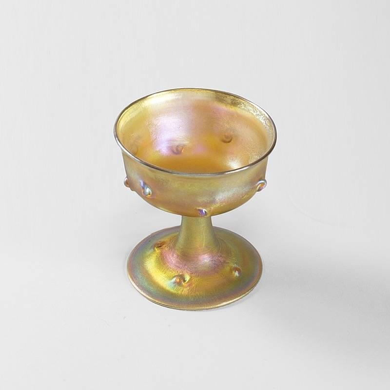 A gold iridescent favrile compote by Louis Comfort Tiffany. The exterior of the compote bowl and its base are decorate with prunts, circa 1900s.

Similar compotes are pictured in: “Louis C. Tiffany: The Collected Works of Robert Koch”, Atglen, PA: