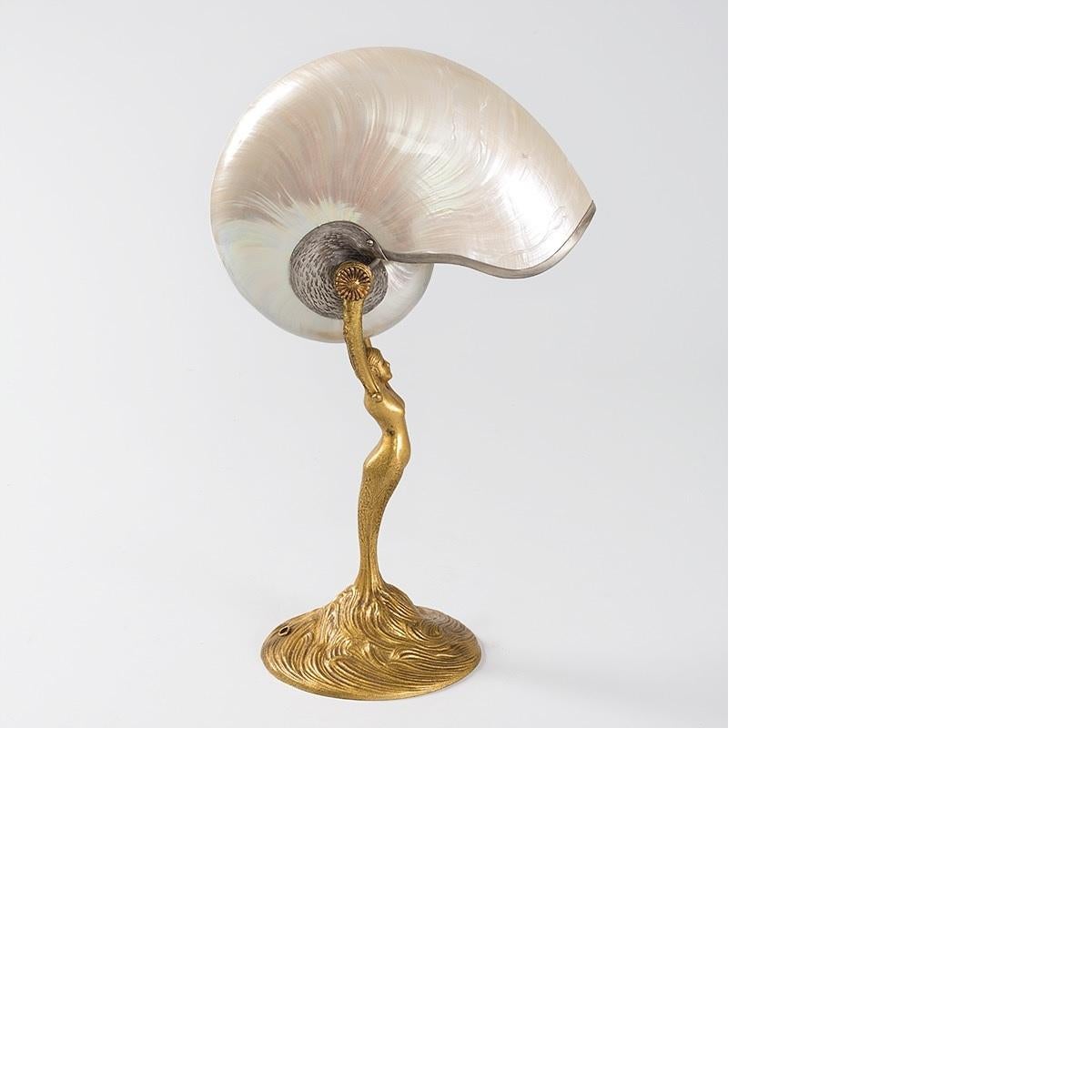 A Tiffany Studios New York “Nautilus” table lamp with a silver-lined nautilus shade suspended within a gilt bronze figural base. The base is in the shape of a mermaid with outstretched arms, emerging from a seascape. When Tiffany conceived the