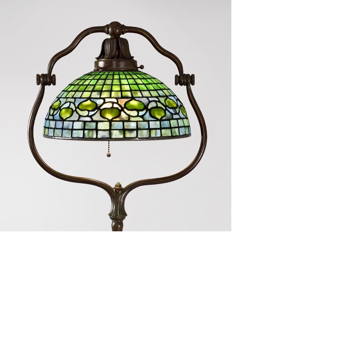 A Tiffany Studios New York “Acorn” bronze and leaded glass floor lamp. The shade feature a yellow acorn leaf and vine motif against a mottled green geometric “brick” ground. The shade is suspended within patinated bronze “Harp” base.

The lamp