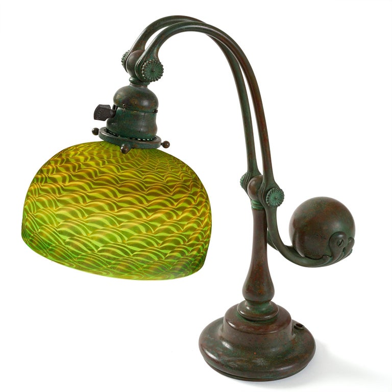 Composed of a damascene favrile domed shade and patinated bronze “Counter Balance” or “Balanced-Weight” base, this Tiffany desk lamp dates from circa 1904. The dichroic green and amber damascene patterned shade is suspended from slender double arms,