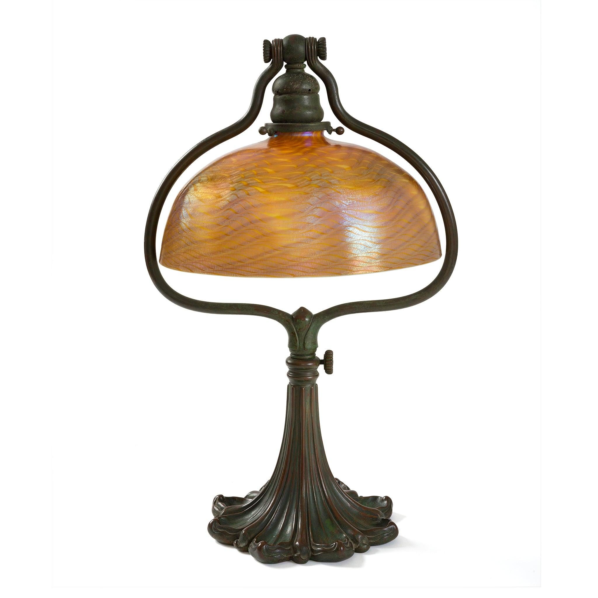 Cette lampe de bureau des studios Tiffany, datant d'environ 1910, présente un abat-jour en verre favrile damasquiné sur une base harpe réglable en bronze patiné. Dans des tons dichroïques ambre-doré et vert, l'abat-jour subtilement irisé est