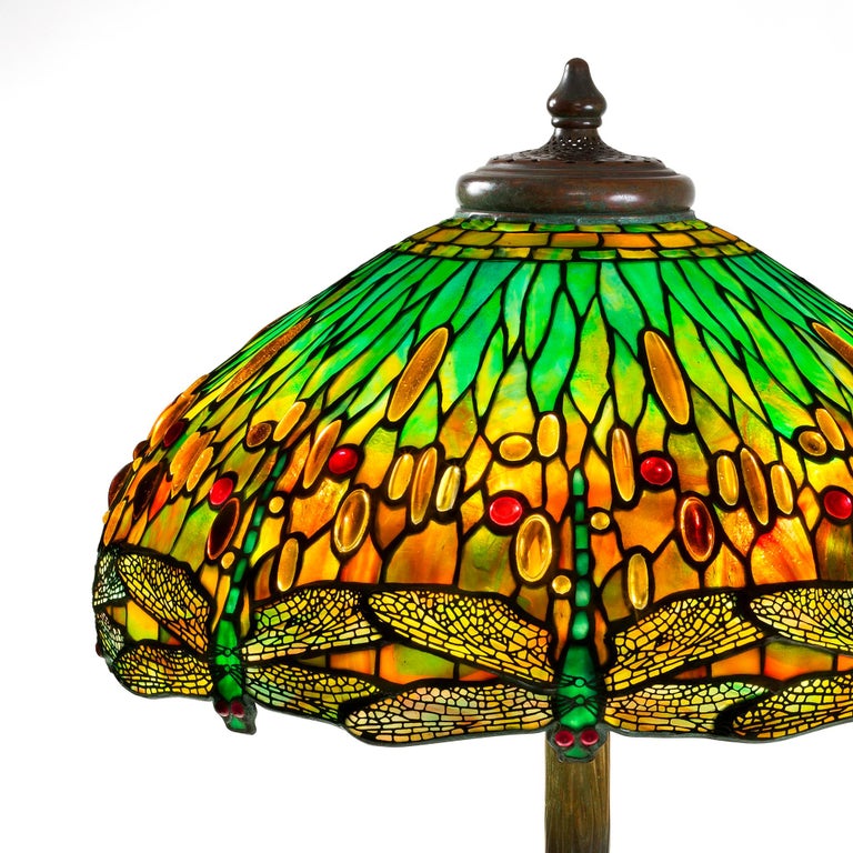 This Tiffany Studios New York glass and bronze “Drophead Dragonfly” table lamp features a leaded glass shade depicting turquoise green dragonflies with mottled turquoise, honey and whiskey colored wings, against a graduated turquoise, green, red and