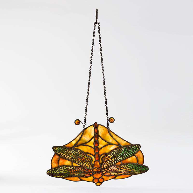 This Tiffany Studios New York “Dragonfly” glass pendant features a single dragonfly, with a reddish-orange body, ombre wings, and topaz jeweled glass eyes, set against a mottled reddish-orange and honey-colored glass ground. Wire mesh is used to
