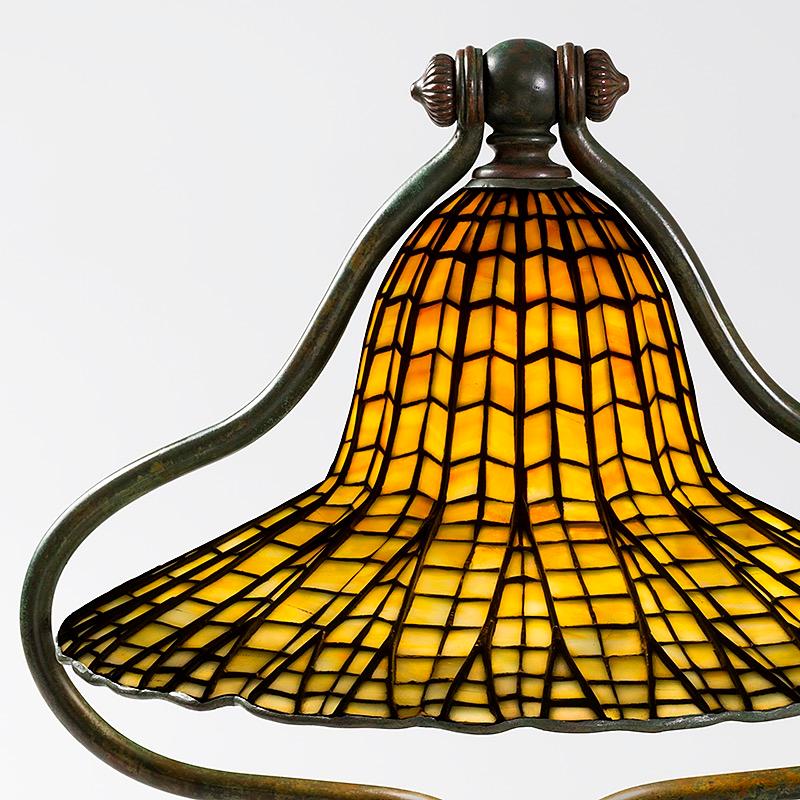 This Tiffany Studios New York leaded glass “Lotus Bell” desk lamp features a beautiful, bell-shaped shade decorated with a cascading geometric design and ombré honey-hued glass tiles. The shade is suspended from a 