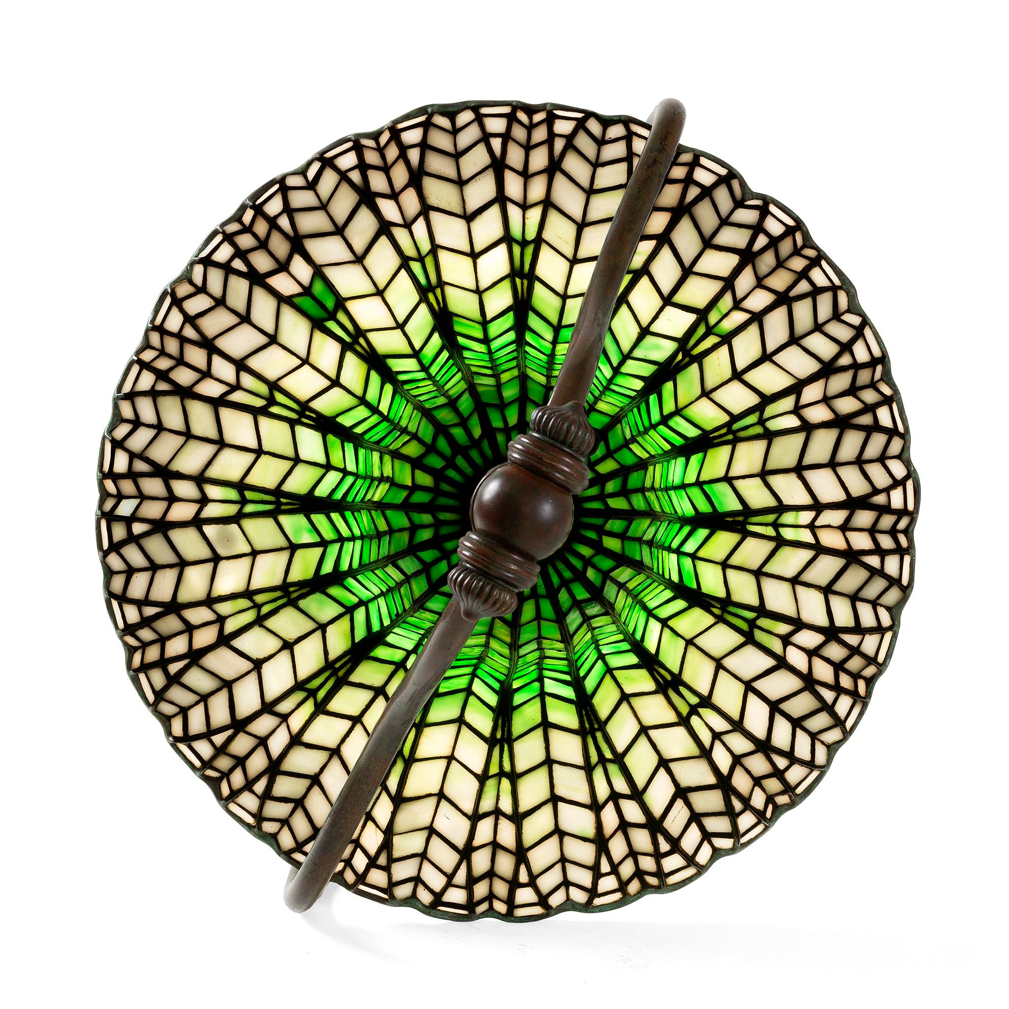 This Tiffany Studios New York glass and bronze “Lotus Bell” table lamp, features a mottled jade green and cream-white leaded glass shade, suspended from a patinated bronze ribbed “Harp” base. The shade bears elaborate veining that required much