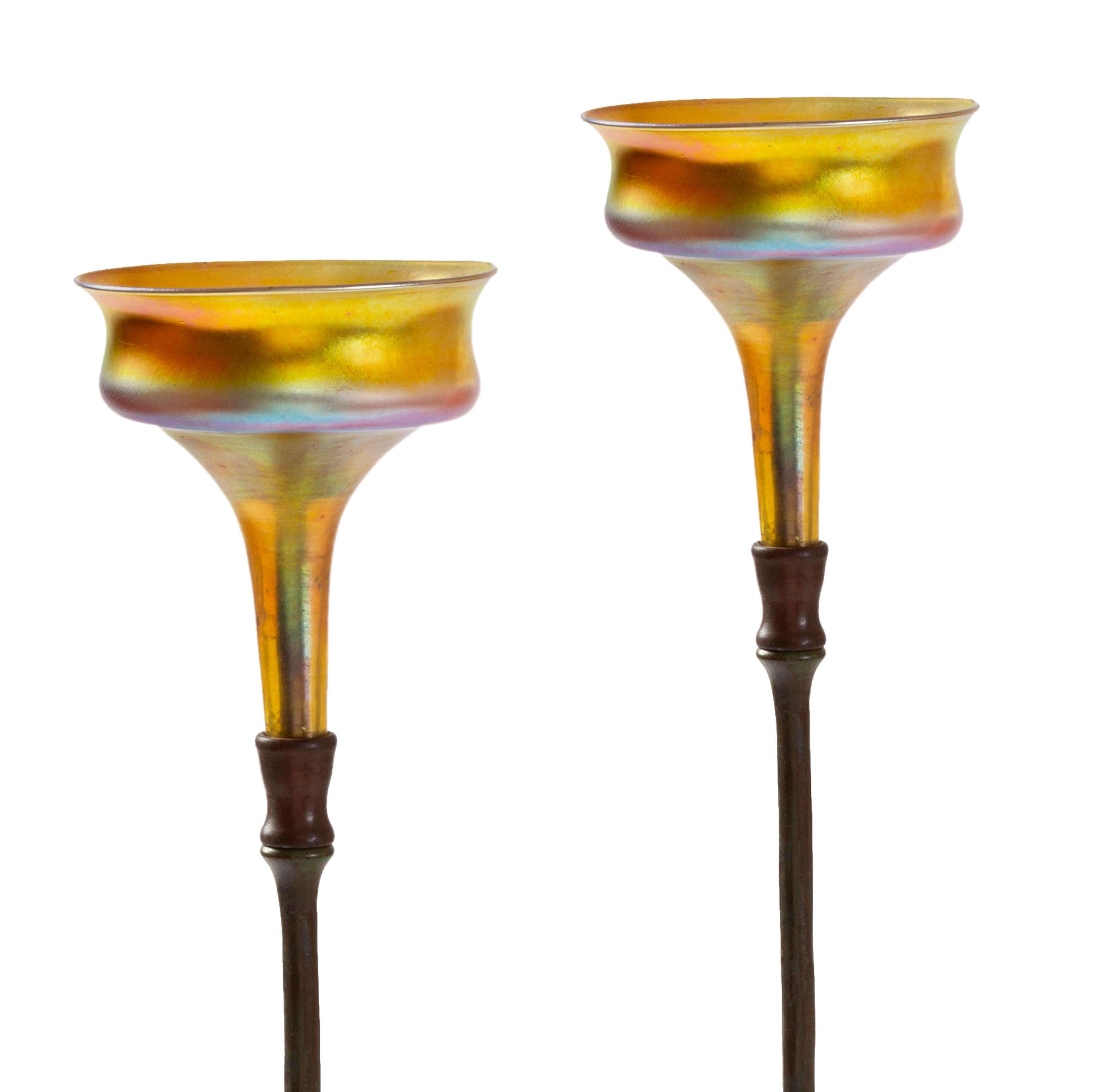 A pair of tall and slender Tiffany Studios New York patinated-bronze candelabra with multihued iridescent blown glass champagne tops. A large disc with rippled edge and swirling texture serves as the foot of the composition, from which a rigid stem,