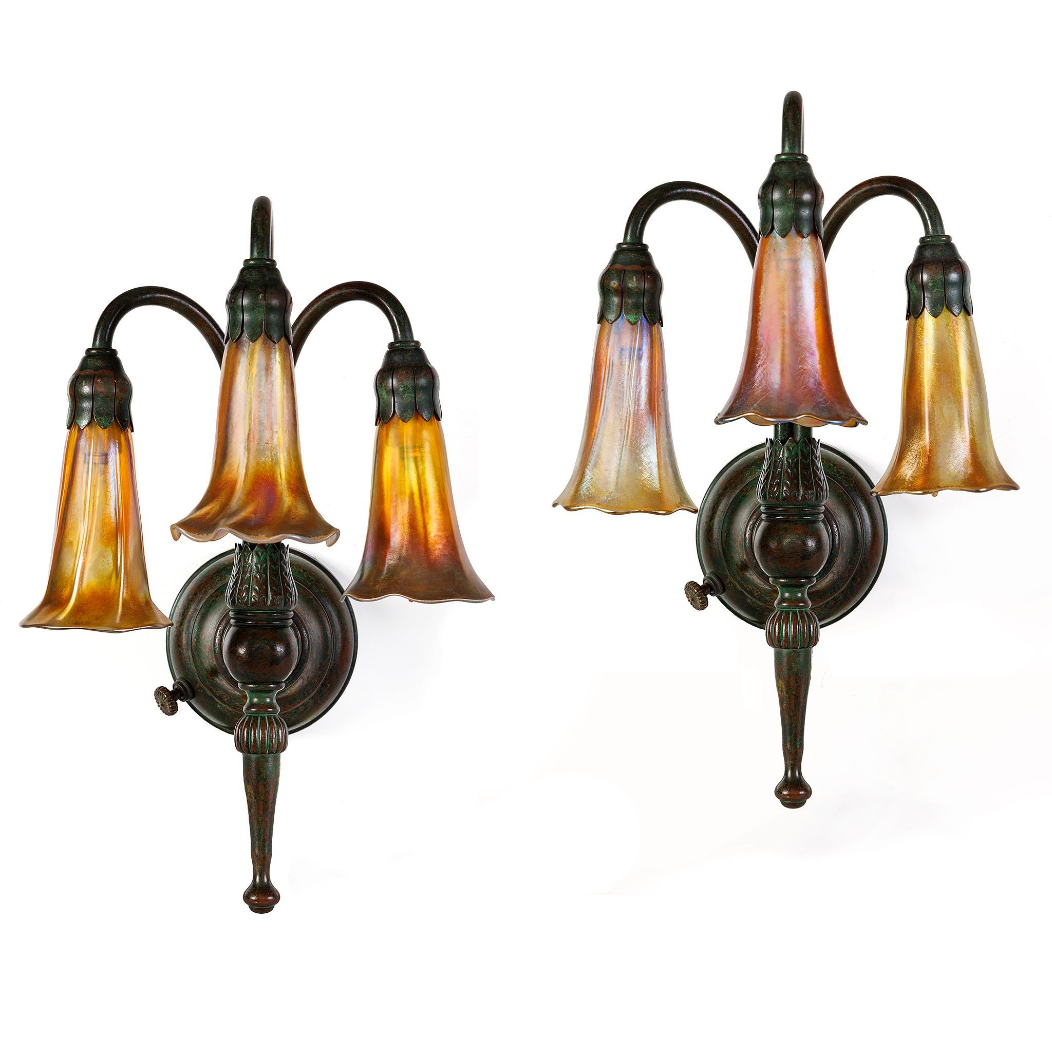A refined interpretation of Tiffany Studios New York’s celebrated Lily design, this matching pair of “Three-Light Lily” wall sconces features golden Favrile glass blossoms that cast warm halos of light across the room. The patinated bronze mounts