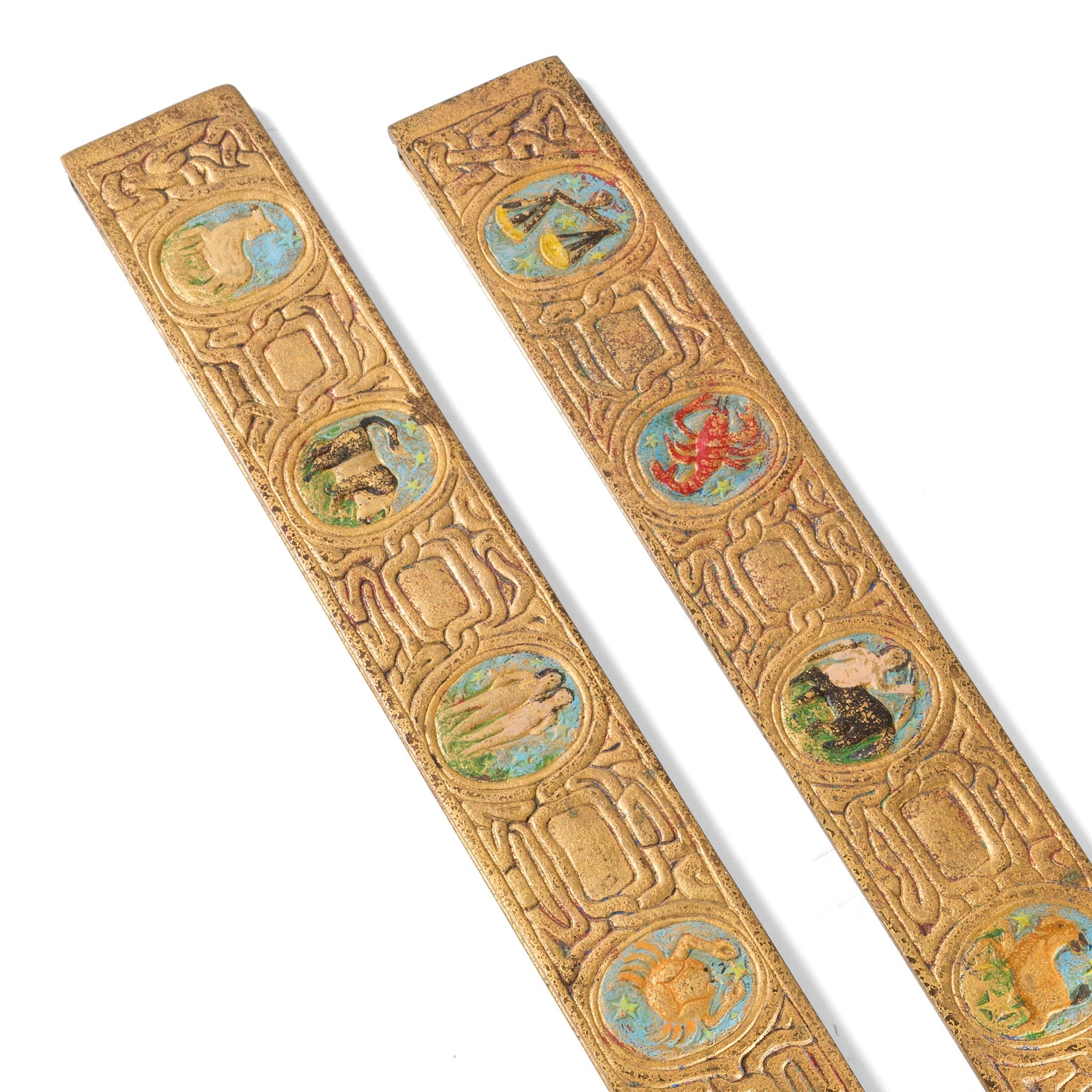 A pair of gilt bronze “Zodiac” desk blotter ends by Tiffany Studios New York. The blotter ends each feature intricate pseudo-Celtic patterning interspersed with circular elements in sky blue, each of which features a stylized depiction of one of the