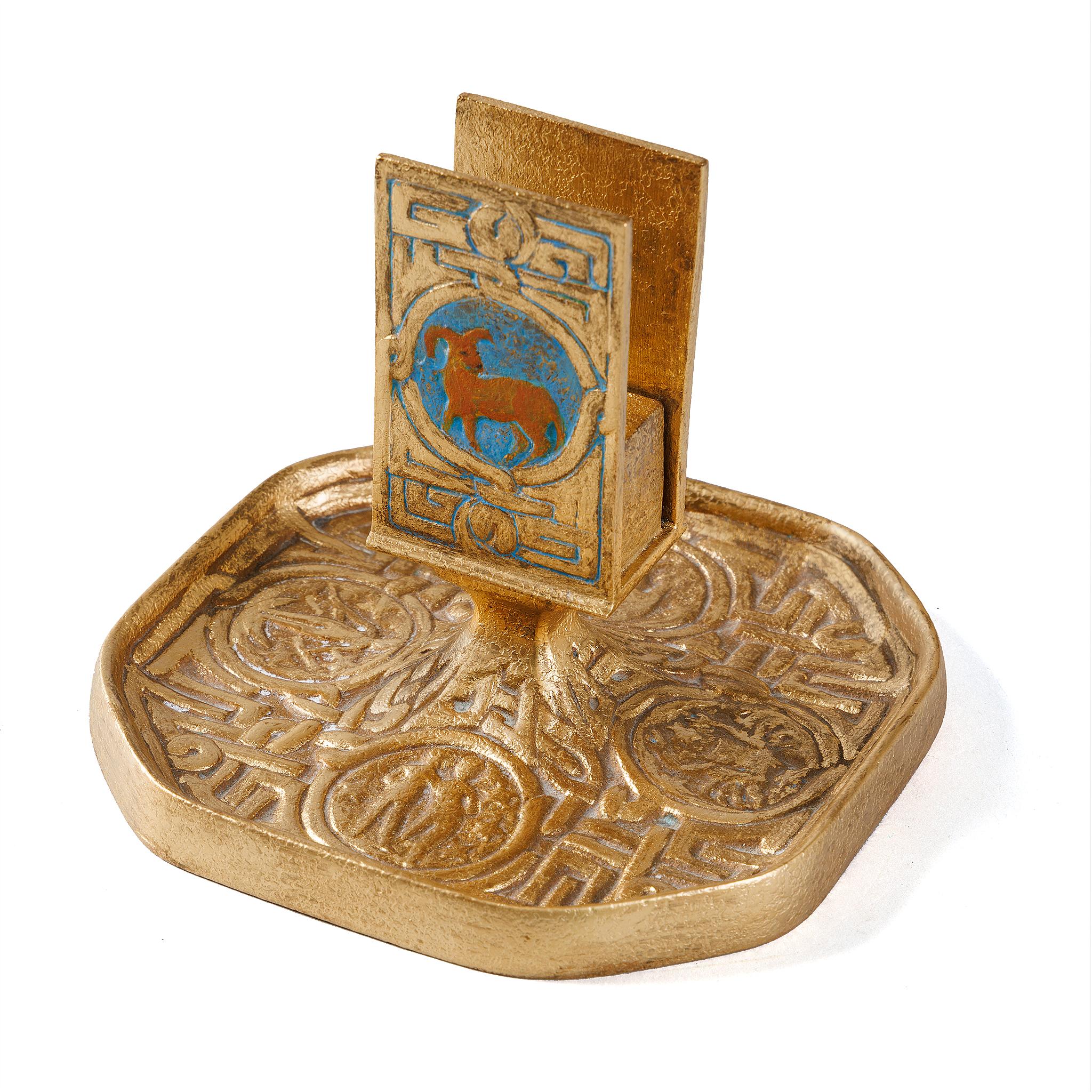 A gilt bronze “Zodiac” match box holder by Tiffany Studios New York. The match box holder features intricate pseudo-Celtic patterning interspersed with circular elements in sky blue, each of which features a stylized depiction of one of the 12 signs