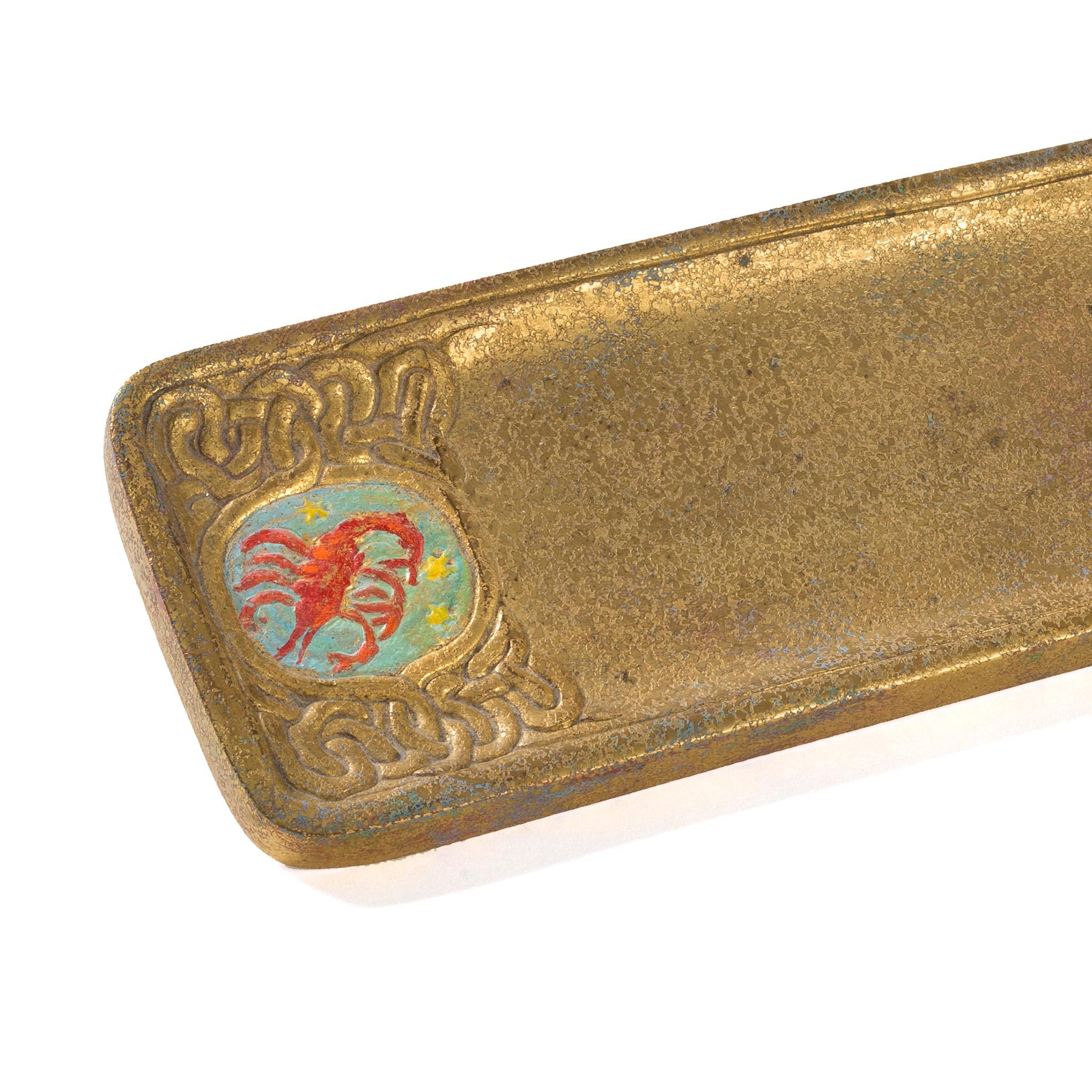 A gilt bronze “Zodiac” pen tray by Tiffany Studios New York. The pen tray features intricate pseudo-Celtic patterning interspersed with circular elements in sky blue, each of which features a stylized depiction of one of the 12 signs of the Zodiac,