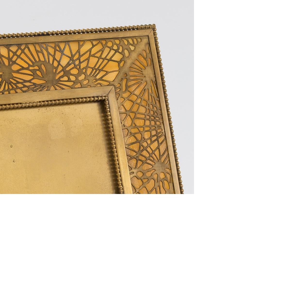 A Tiffany Studios New York gilt bronze and Favrile glass picture frame in the “Pine Needle” pattern with an square center and beaded trim, mottled amber and opaque colored glass, circa 1900.

A similar picture frame is pictured in: Tiffany Desk